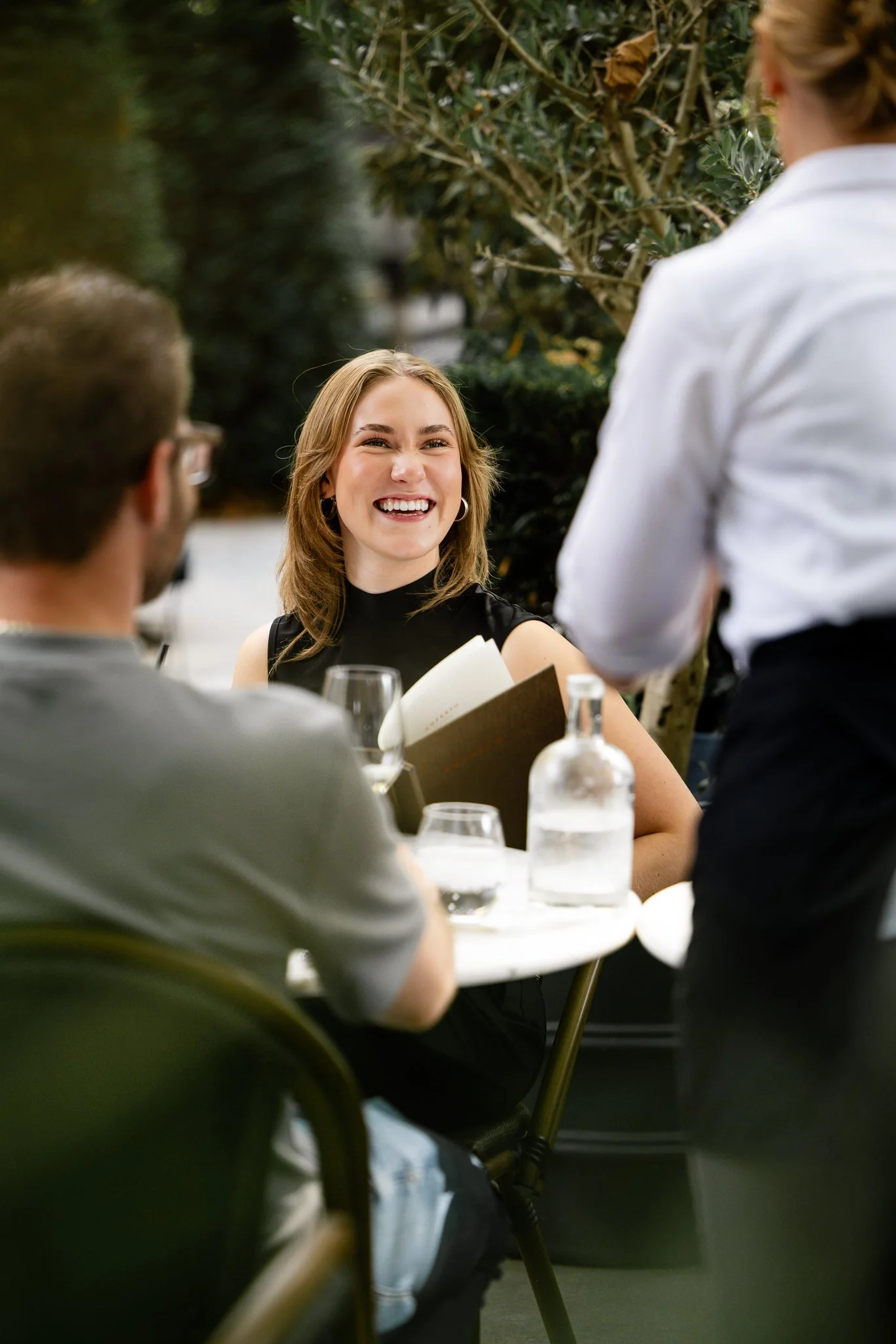 Een vrouw die lacht en een menukaart vasthoudt aan een tafel in een restaurant, terwijl een ober haar bedient. Op de achtergrond groene planten.