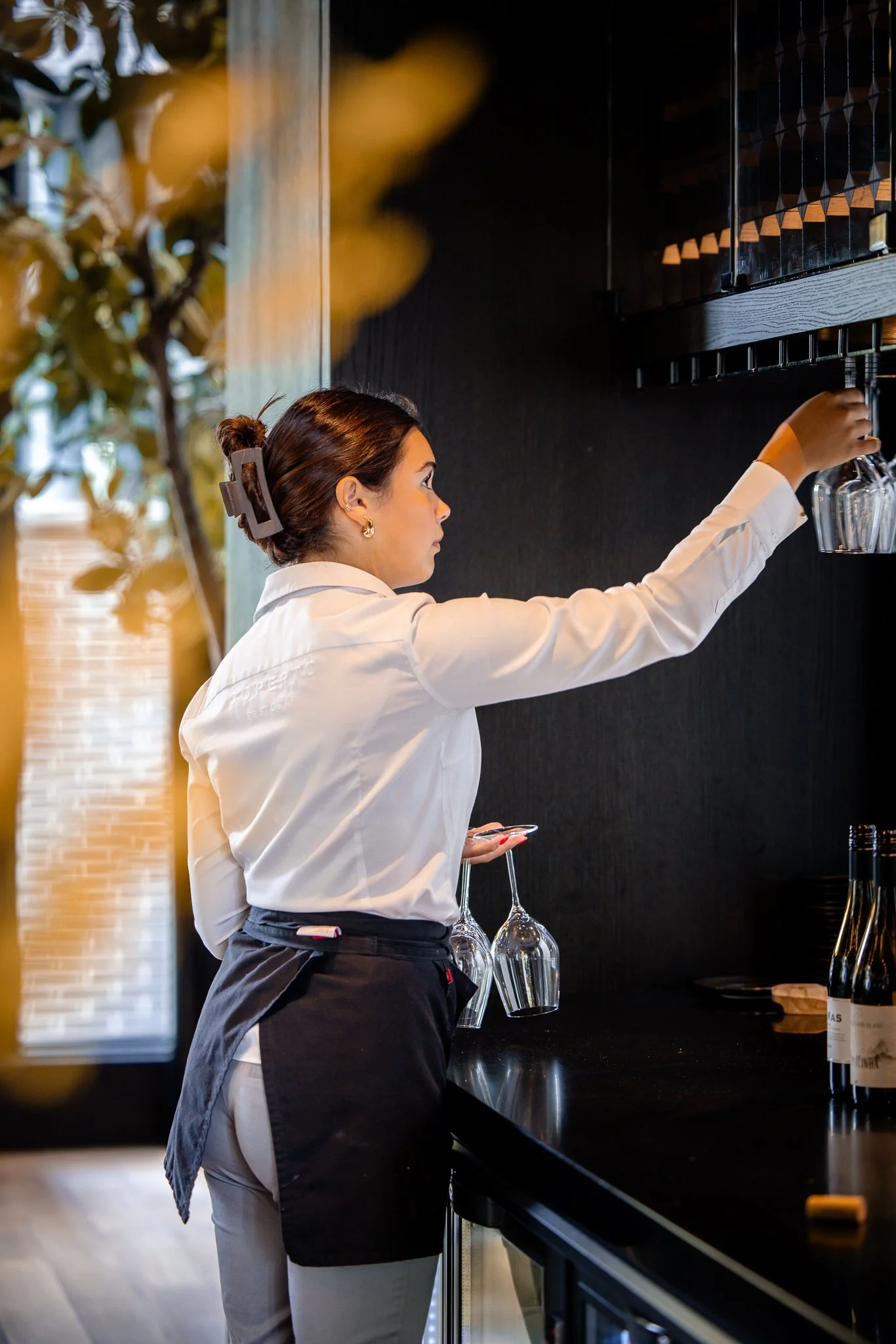 Een serveerster met een witte blouse en een zwarte schort die glazen vast heeft in een restaurant, met borrelglazen en een wijnfles op de counter.