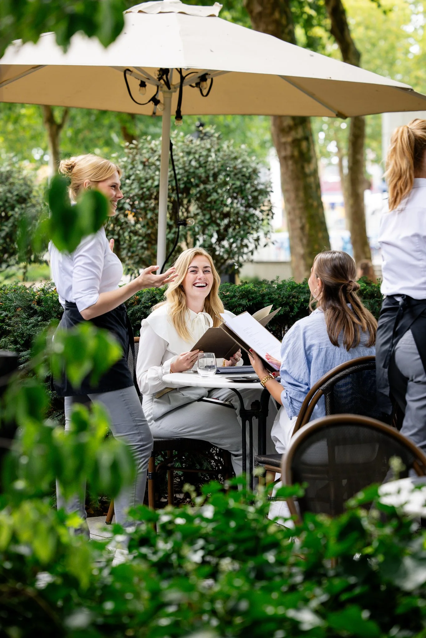 Een groep vrouwen zit en staat buiten onder een grote parasol, omgeven door groen, en lijken een gesprek te voeren in een tuin of op een terras.