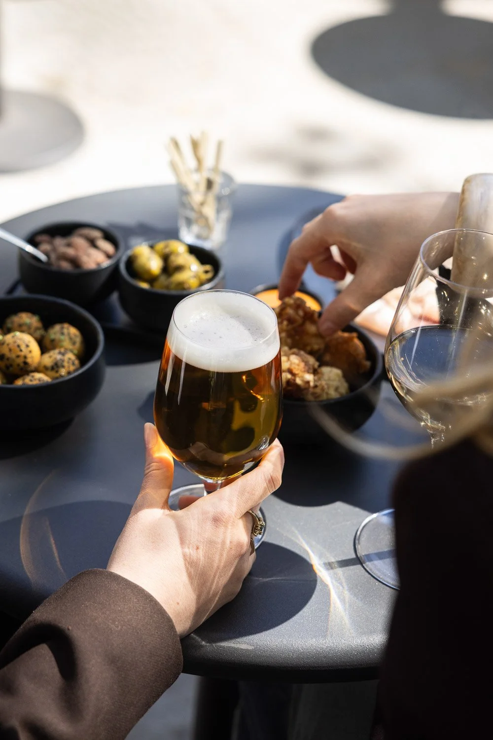 Mensen drinken bier en eten tapas op een buitentafel met verschillende hapjes en glazen wijn.
