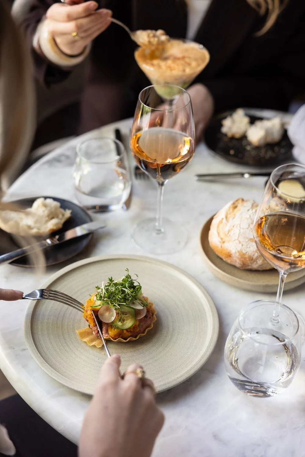 Gasten genieten van een diner met wit wijnglas en hapje op bord, met brood en drankjes op de tafel.
