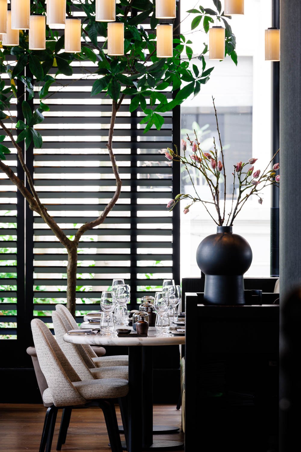 Een decoratief interieur met een grote plant met groene bladeren, een tafel met glazen en bestek, en een grote zwarte vaas met takken met roze bloemen, binnen en met een raam op de achtergrond.