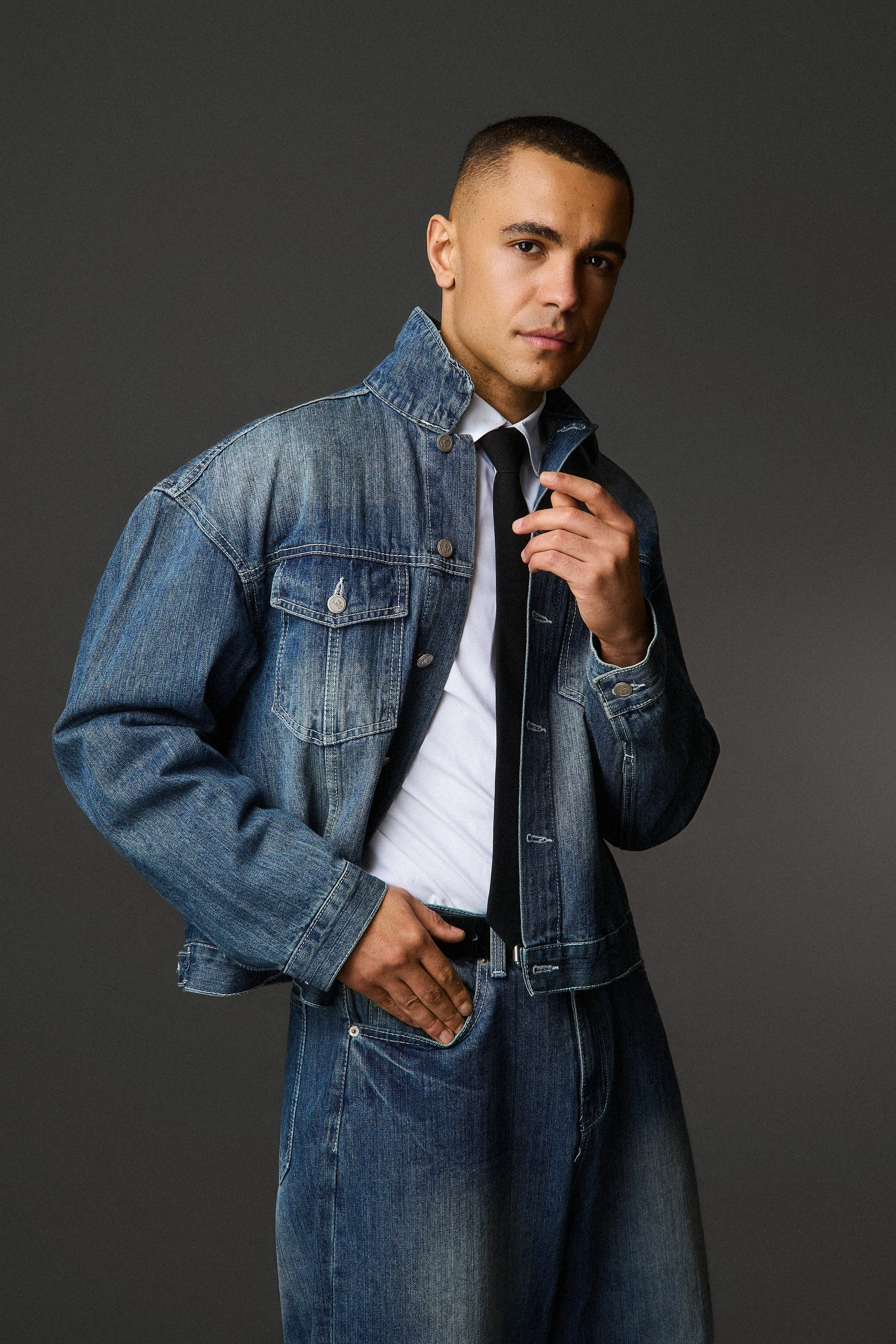 A young man with short hair, wearing a denim jacket over a white shirt and black tie, posing against a dark gray background.