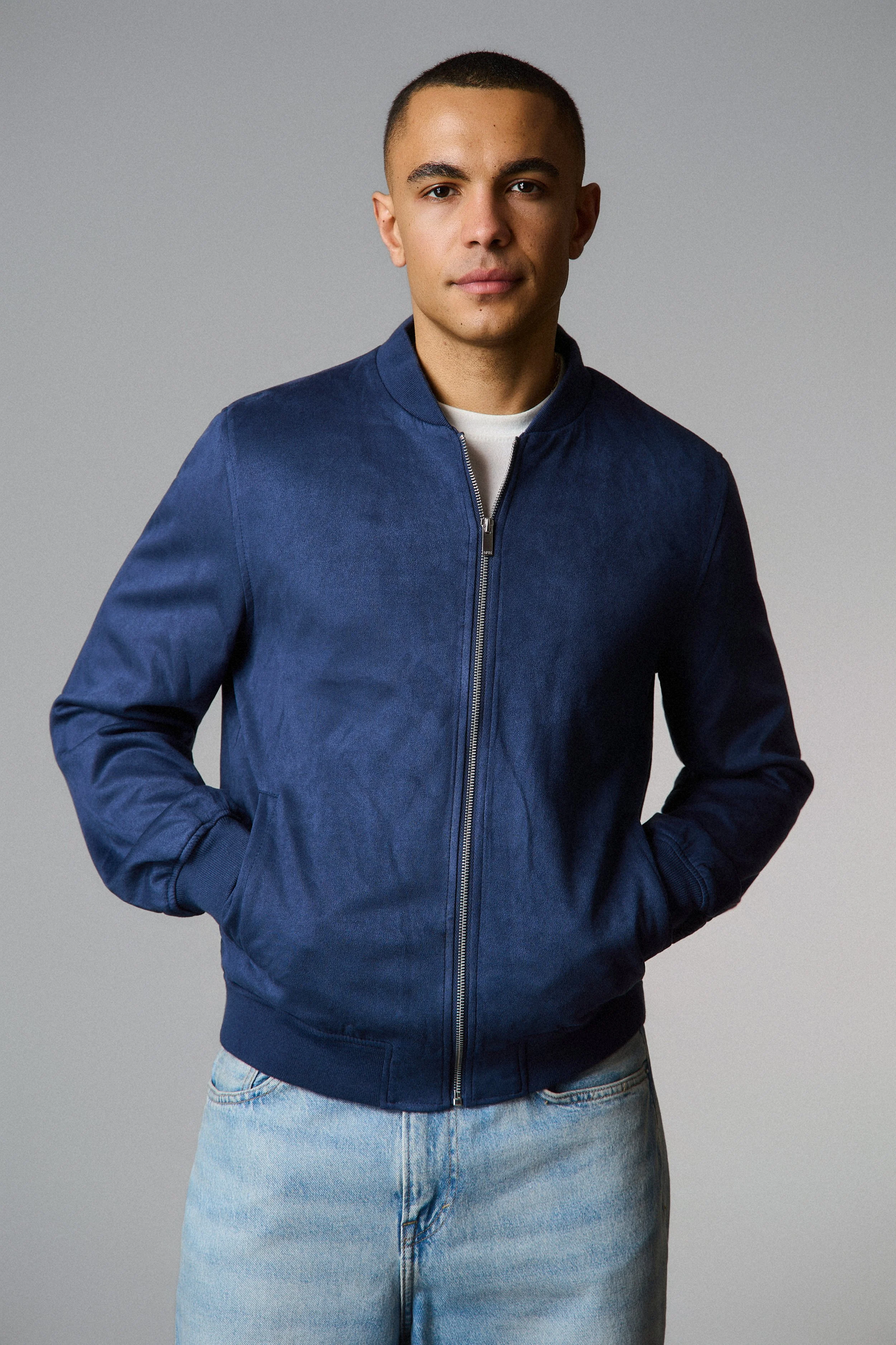 A young man with short dark hair and tan skin, wearing a navy blue zip-up jacket over a white T-shirt, and light blue jeans, standing against a plain gray background.