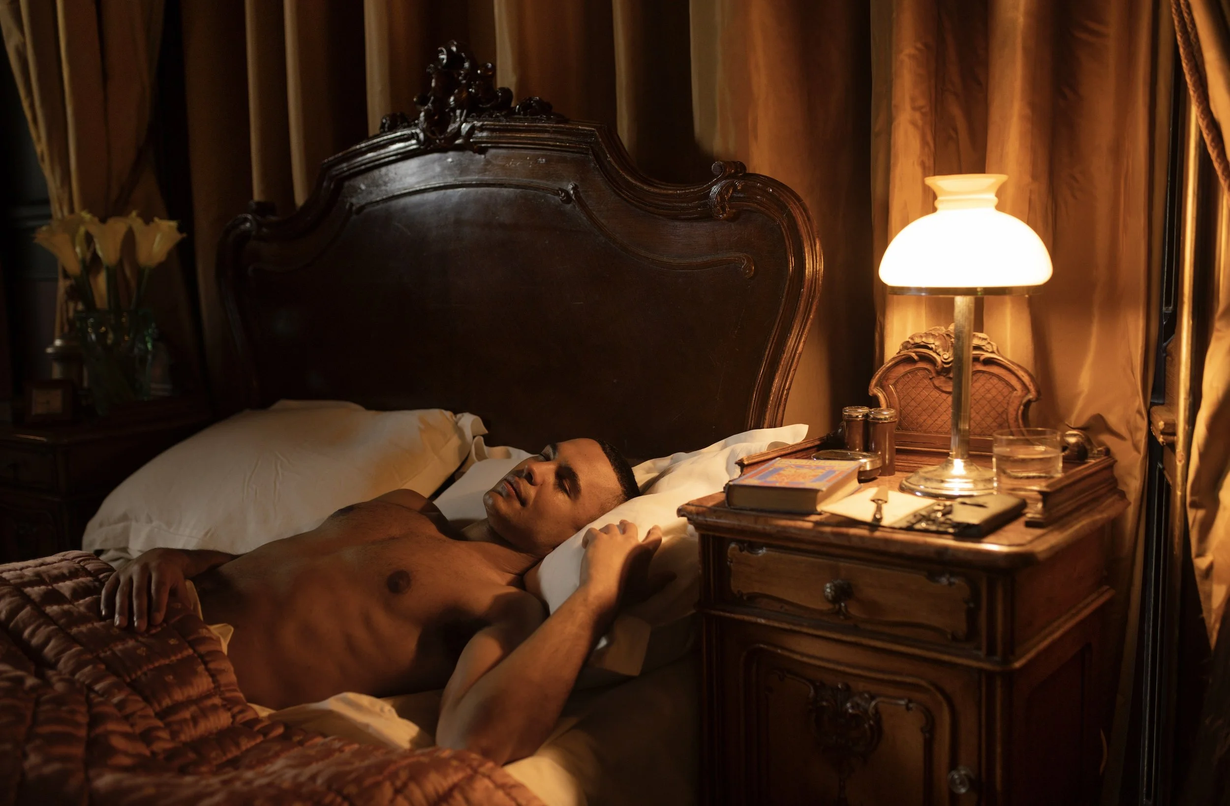 A man is sleeping on a bed in a dimly lit room with a vintage wooden headboard, next to a nightstand with a lamp, books, and glasses. There are curtains and flowers in the background.