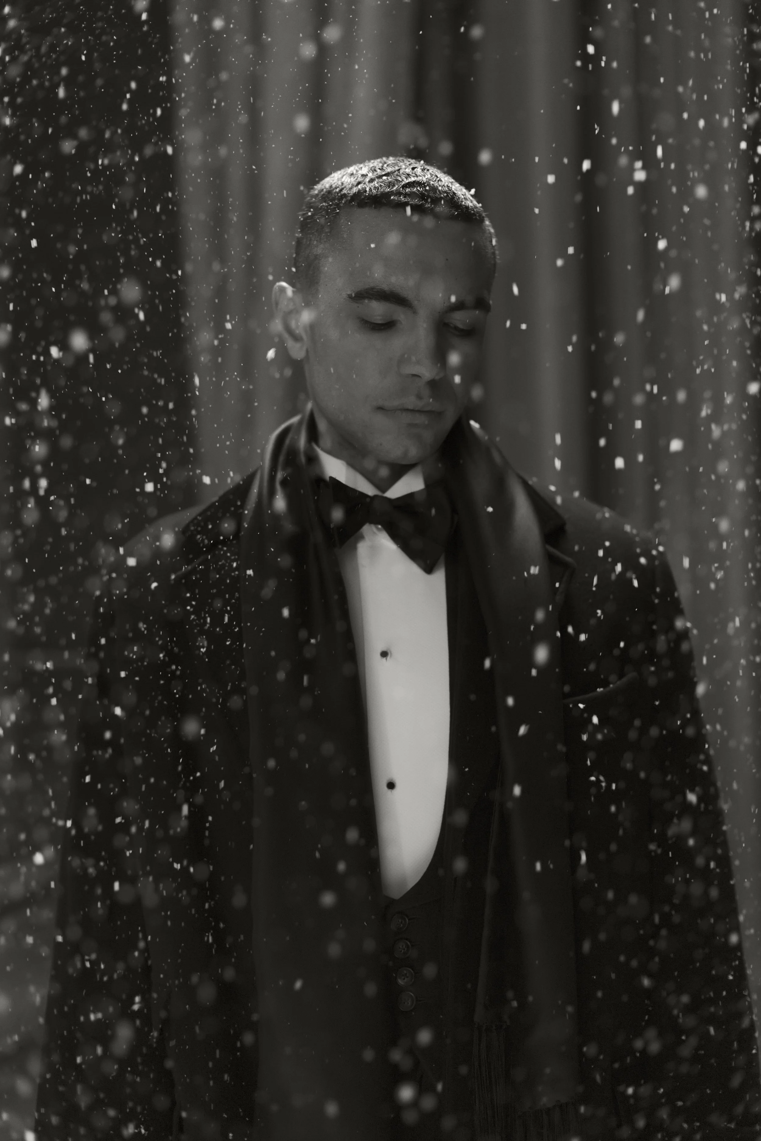 A man dressed in a tuxedo and bow tie with eyes closed, surrounded by falling snow or snow-like particles, with a dark and blurred background.