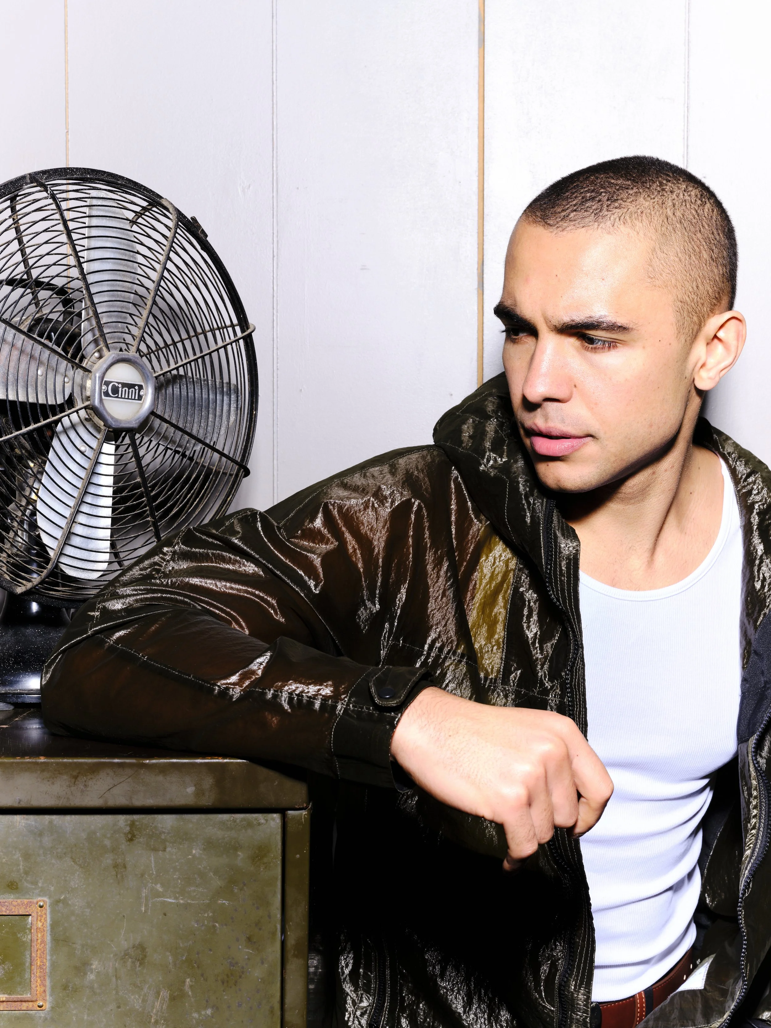 A young man with a shaved head wearing a white t-shirt and a dark shiny jacket, sitting next to an industrial fan inside a room with white paneled walls.