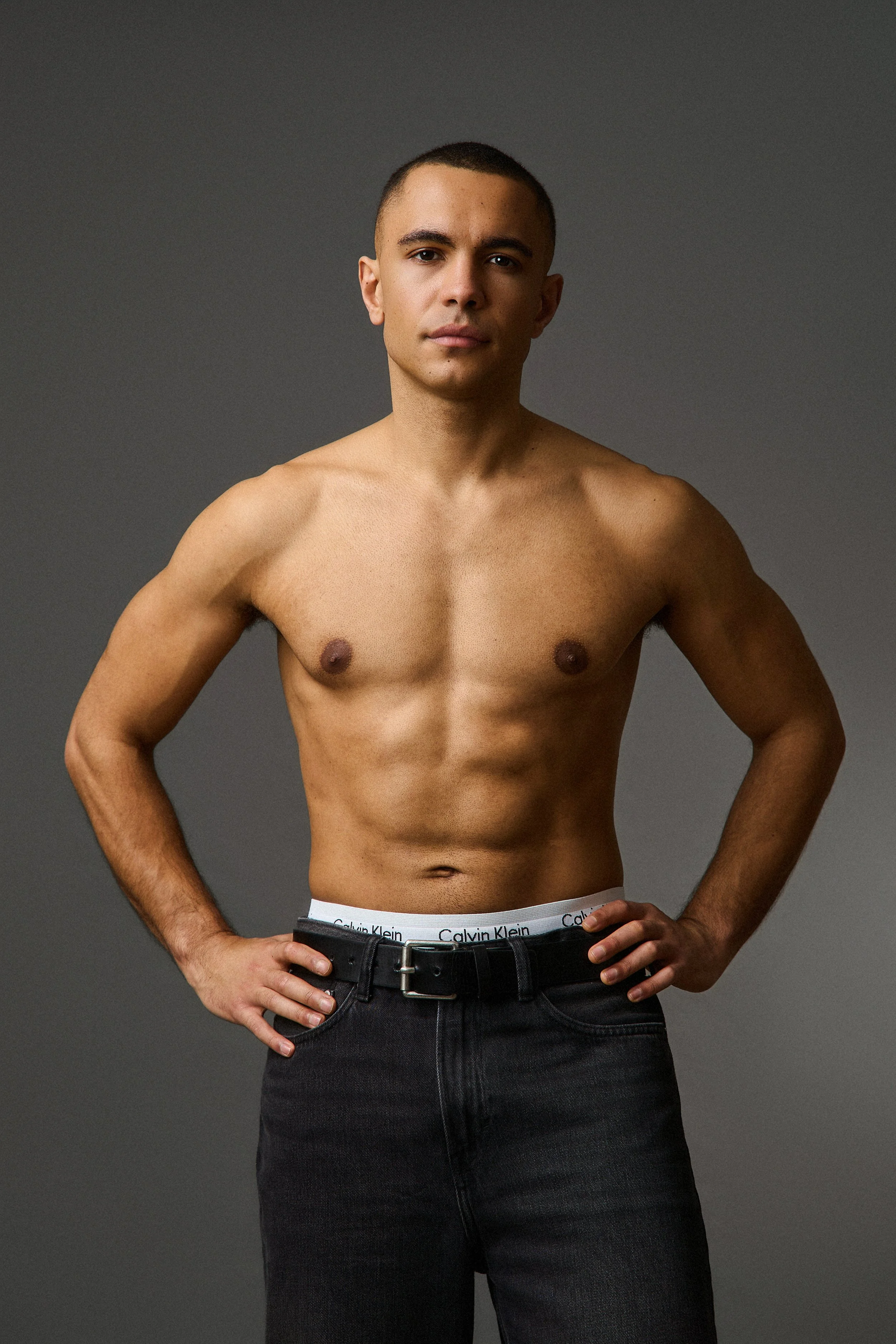 A shirtless young man with short dark hair, standing with his hands on his hips against a gray background, wearing black jeans and white Calvin Klein underwear.