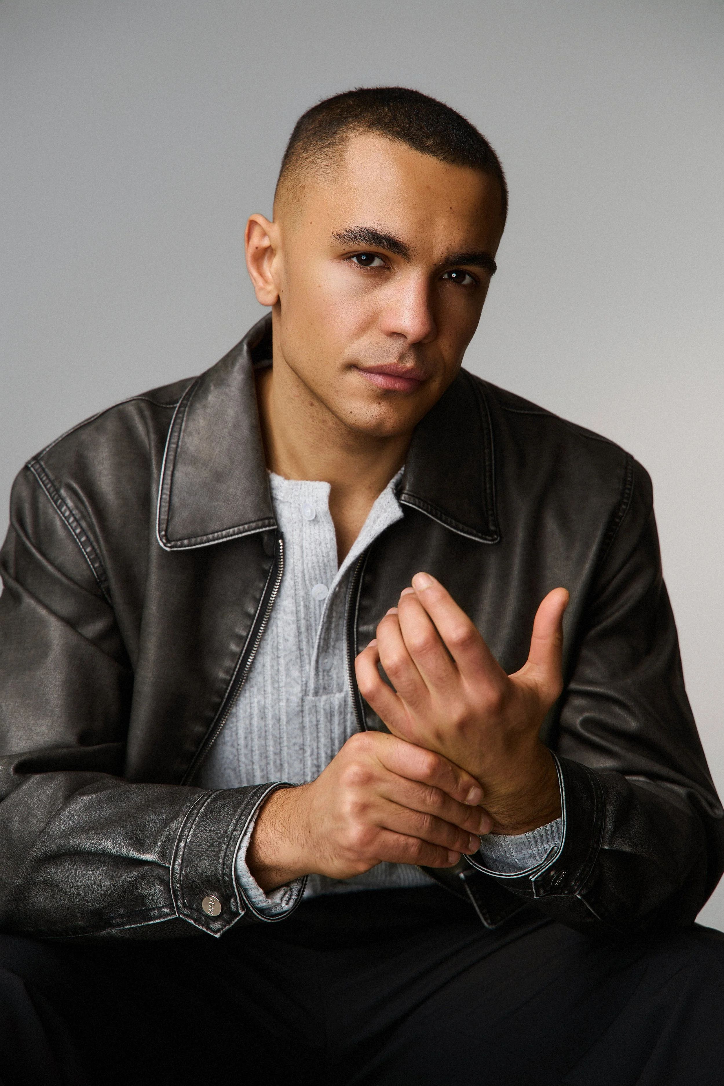 A young man with short dark hair, wearing a black leather jacket over a gray sweater, is looking directly at the camera with a neutral expression. His hands are positioned near his chest, with one hand gripping the other.
