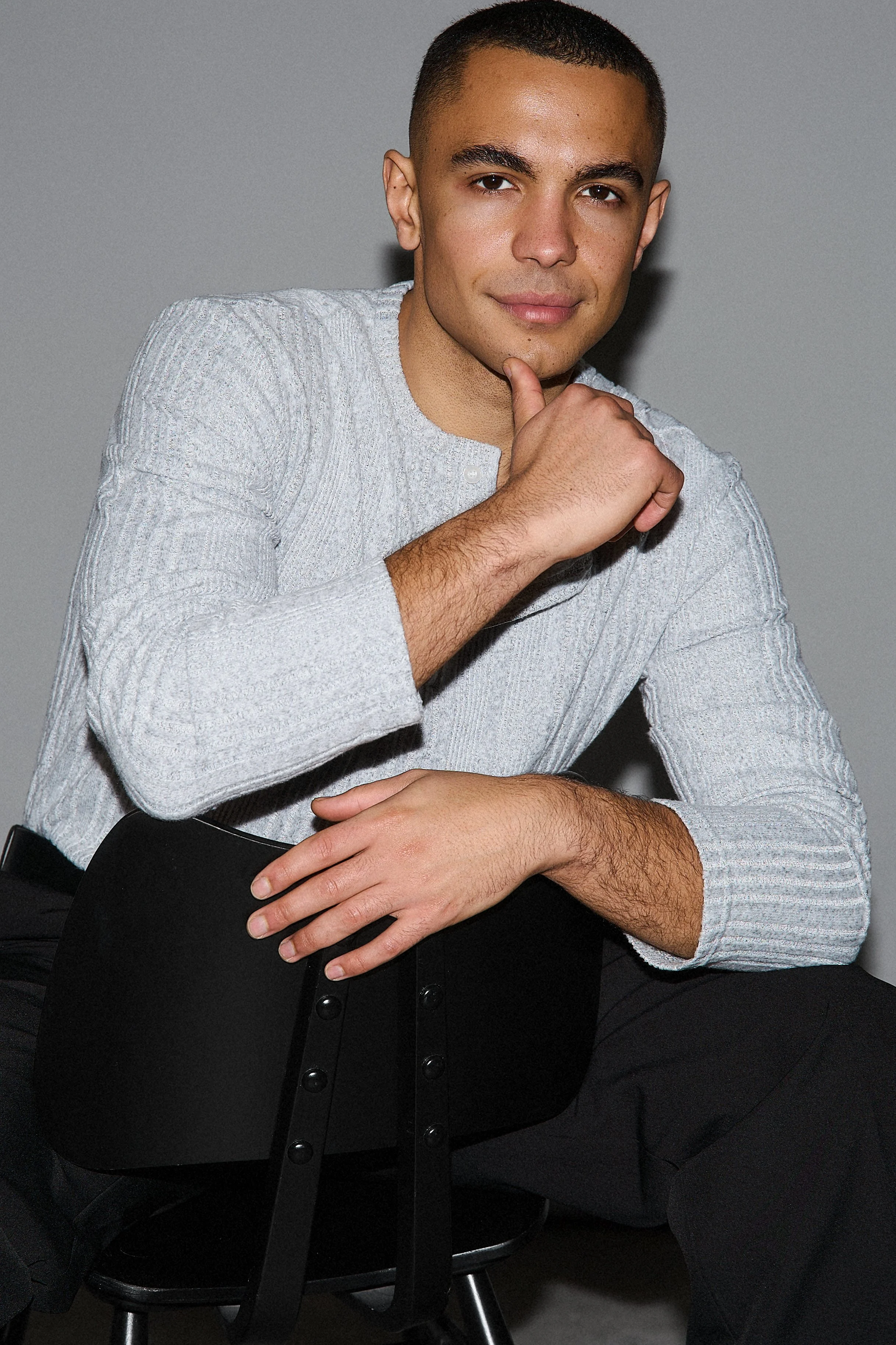 A young man with short dark hair and tan skin poses sitting on a black chair, wearing a light gray textured long-sleeve shirt and black pants. He rests his chin on his right hand and his left arm is crossed over his lap against a plain gray background.