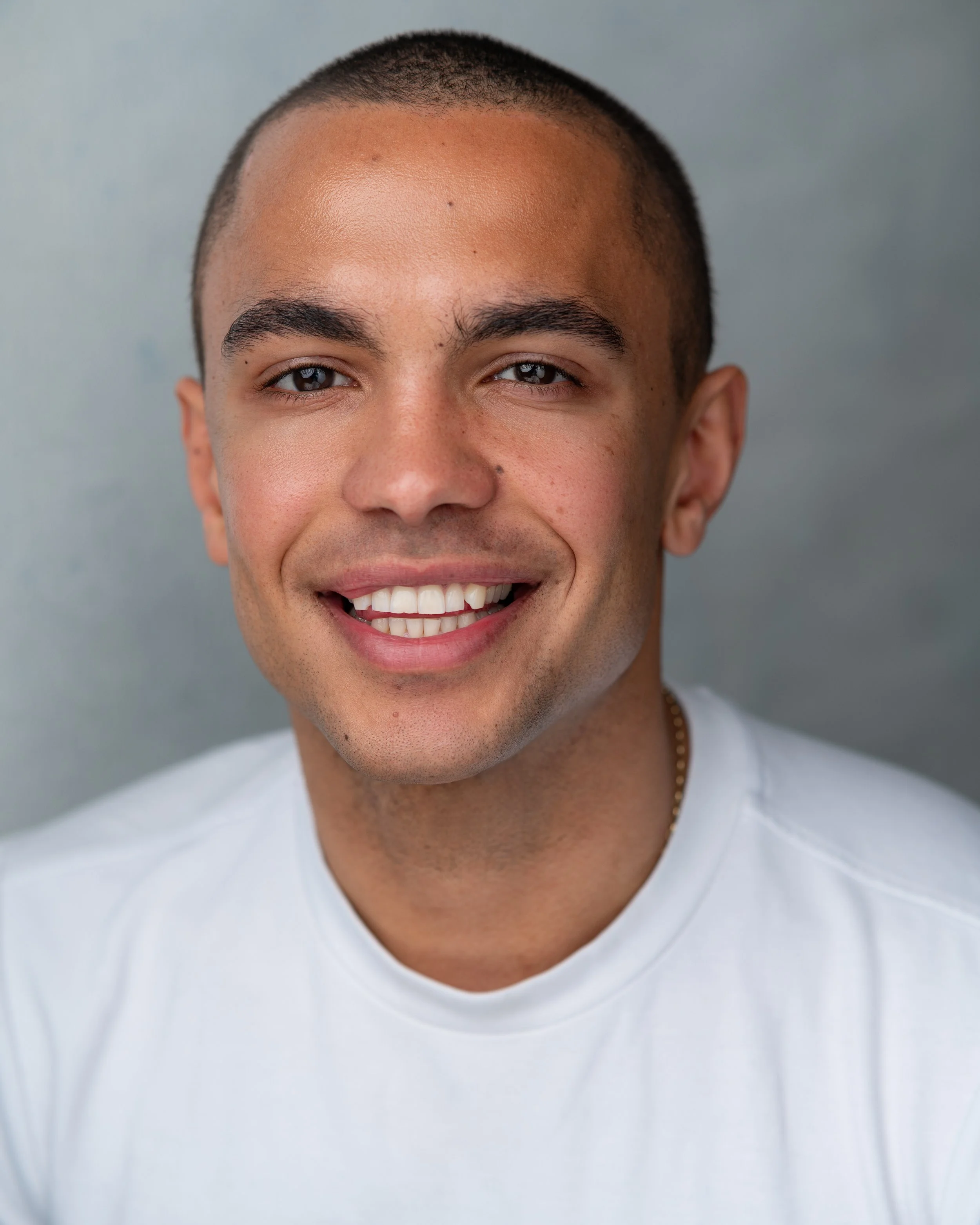 Close-up of smiling young man with short hair, wearing a white shirt, against a plain gray background.