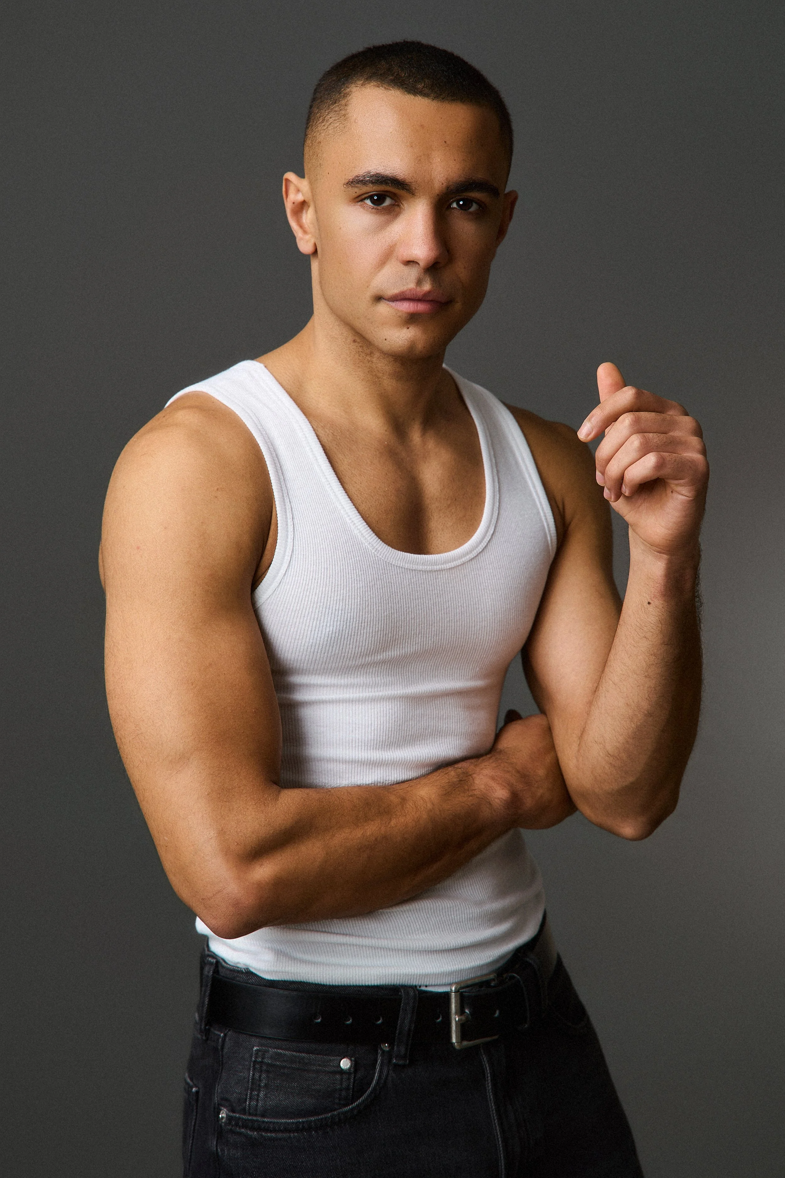 A young man with short dark hair wearing a white tank top and black pants standing against a gray background, posing with his right arm crossed and left hand touching his chin.