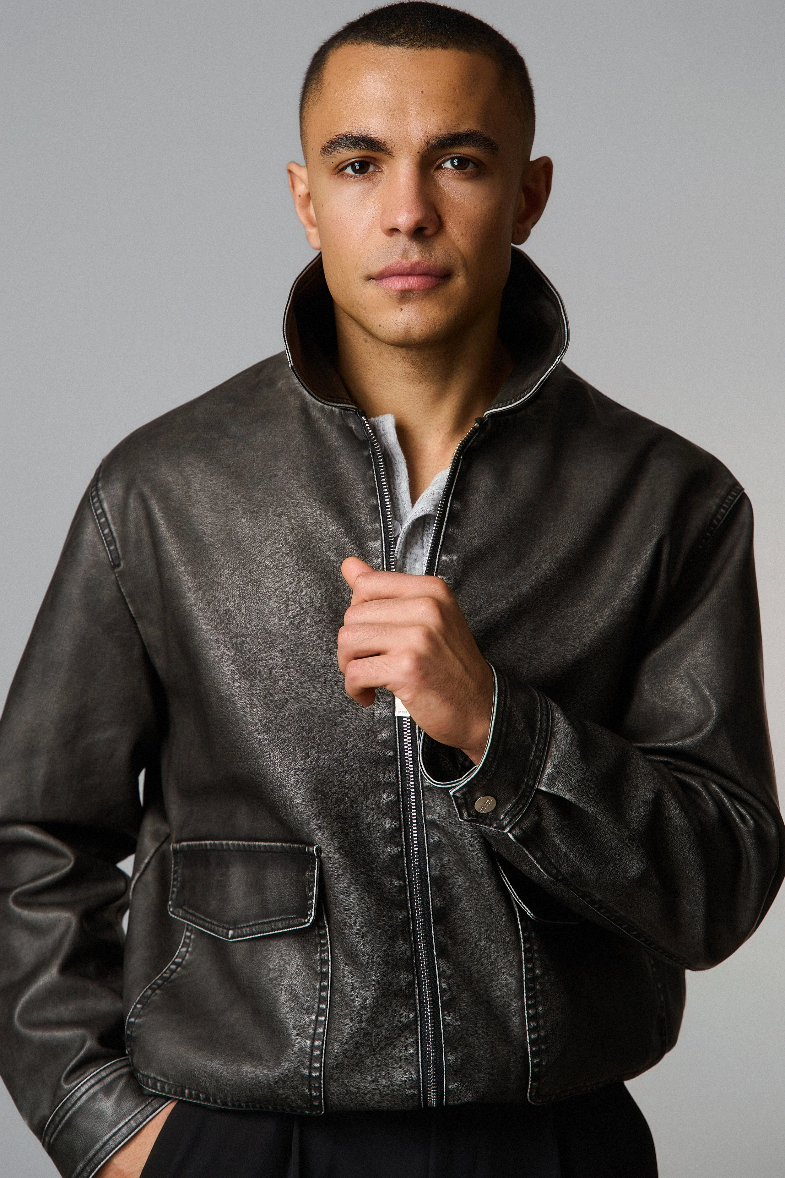 A young man with short dark hair and light skin, wearing a black leather jacket with a popped collar over a white shirt, standing against a plain gray background.