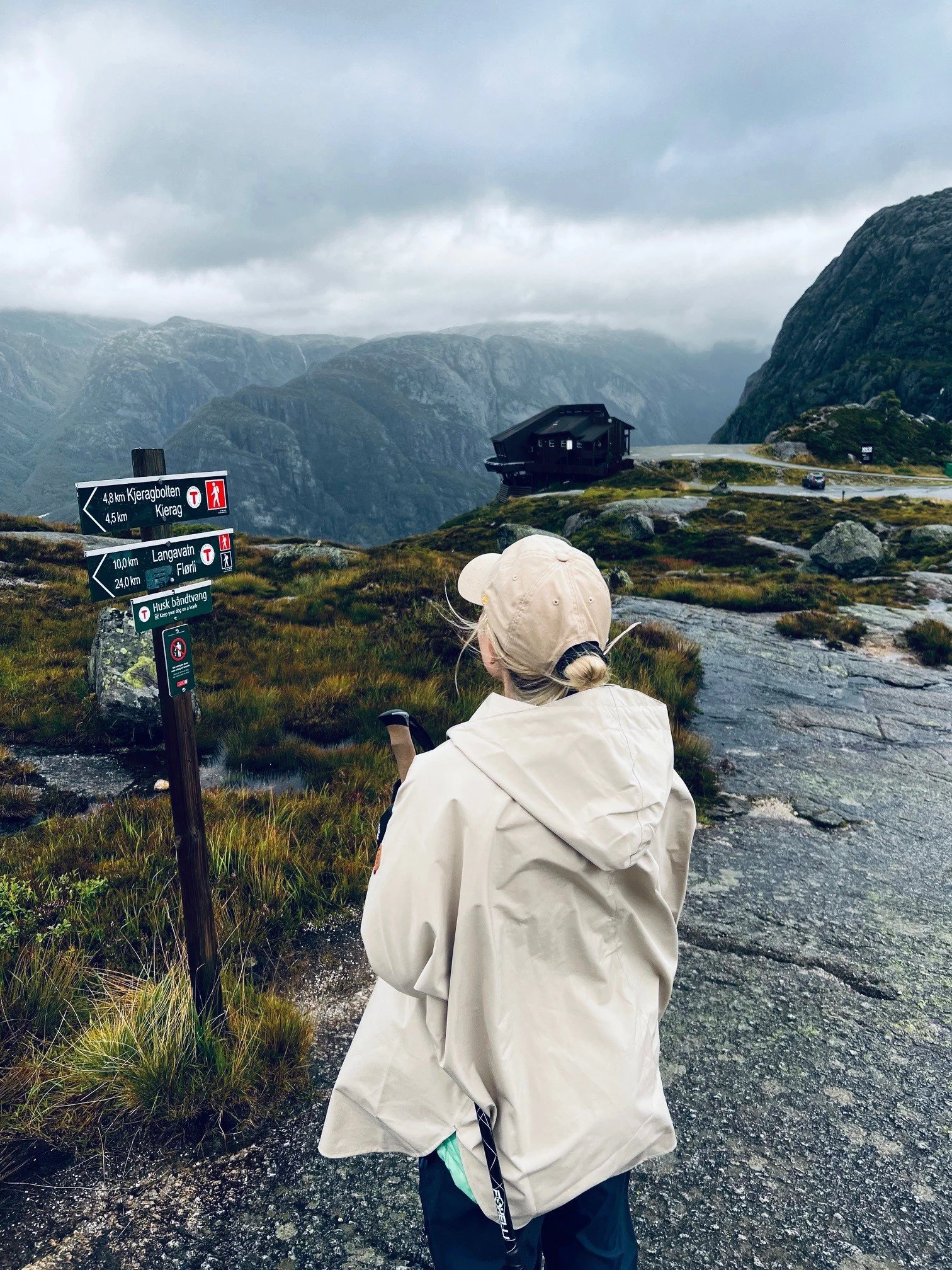 A person wearing a light-colored jacket and a baseball cap is standing on a rocky, grassy mountain trail, looking at a signpost that indicates distances to nearby locations. There are mountains and cloudy skies in the background.