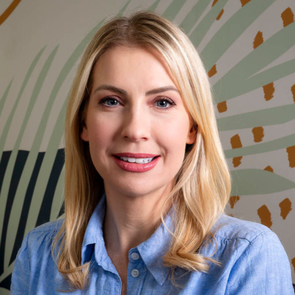 A woman with blonde hair smiling, wearing a light blue button-up shirt, standing in front of a leafy green and tan patterned background.