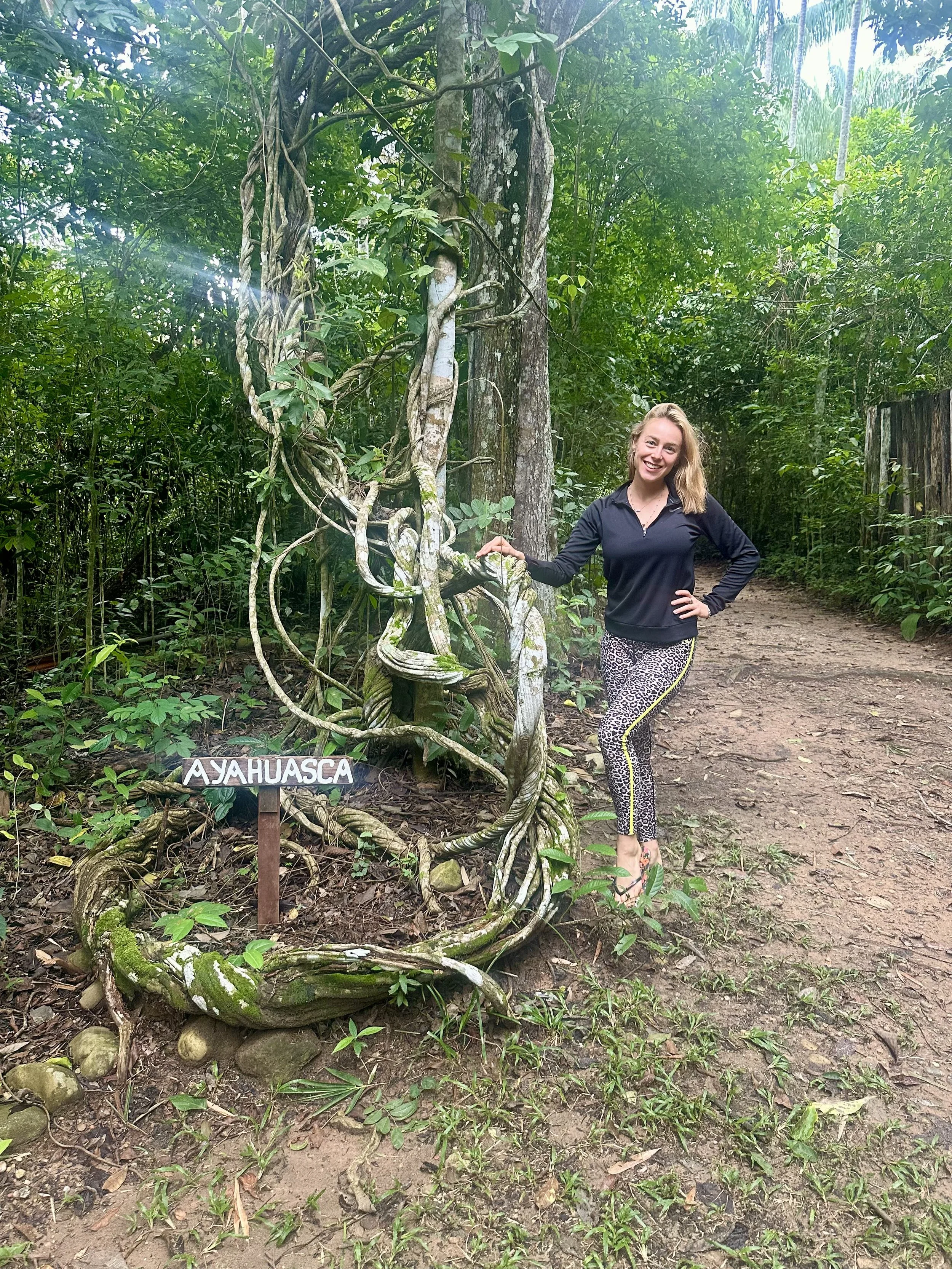 A woman standing on a dirt path in a lush green jungle, smiling and holding a vine. A small sign with the word 'AYAHUASCA' is in front of the vine. The woman has blonde hair, is wearing a dark long-sleeve top and leopard print pants, and is barefoot.