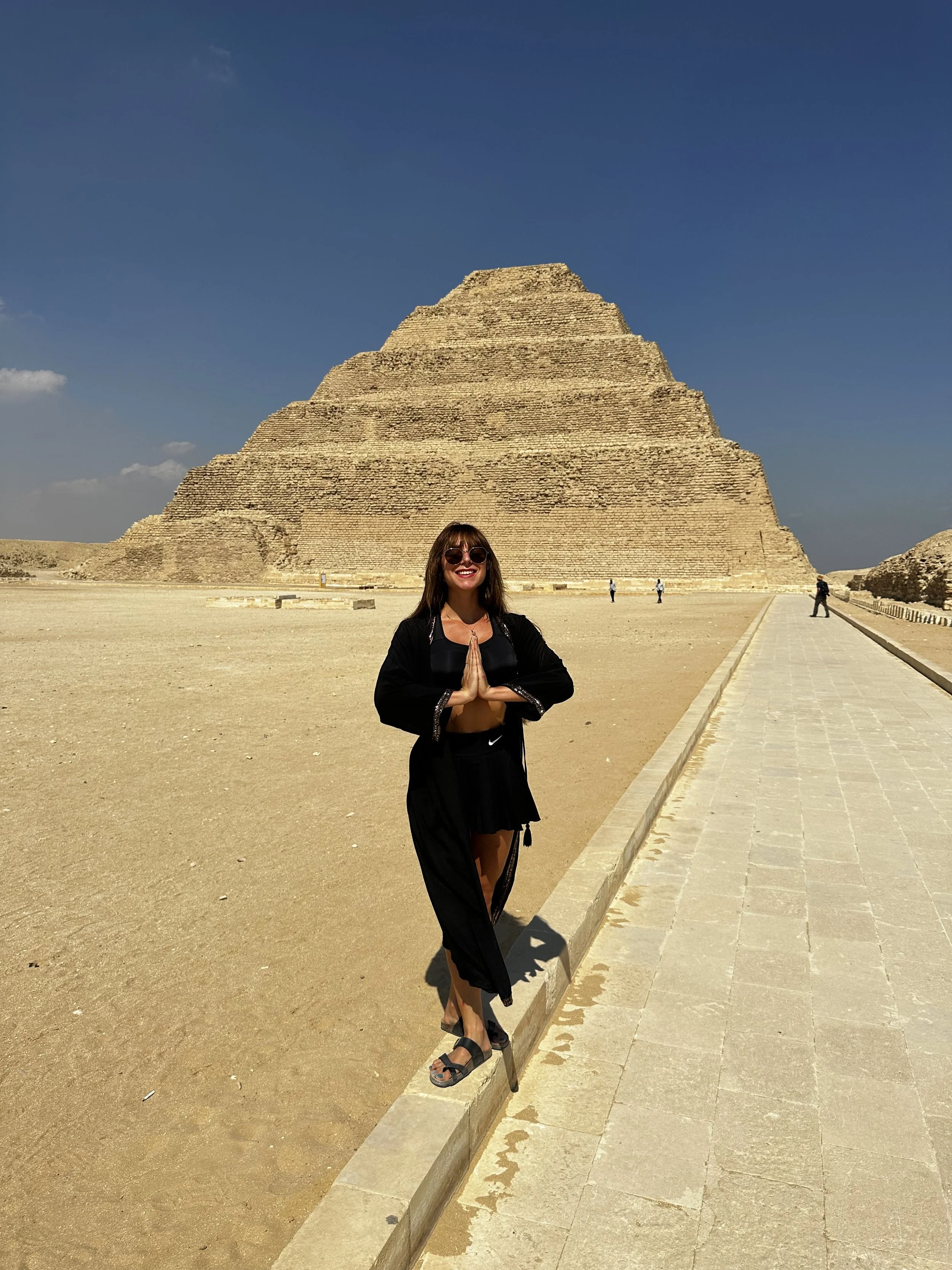 A woman with sunglasses smiling, standing on a small stone ledge, with the step pyramid of Djoser in the background under a clear blue sky.