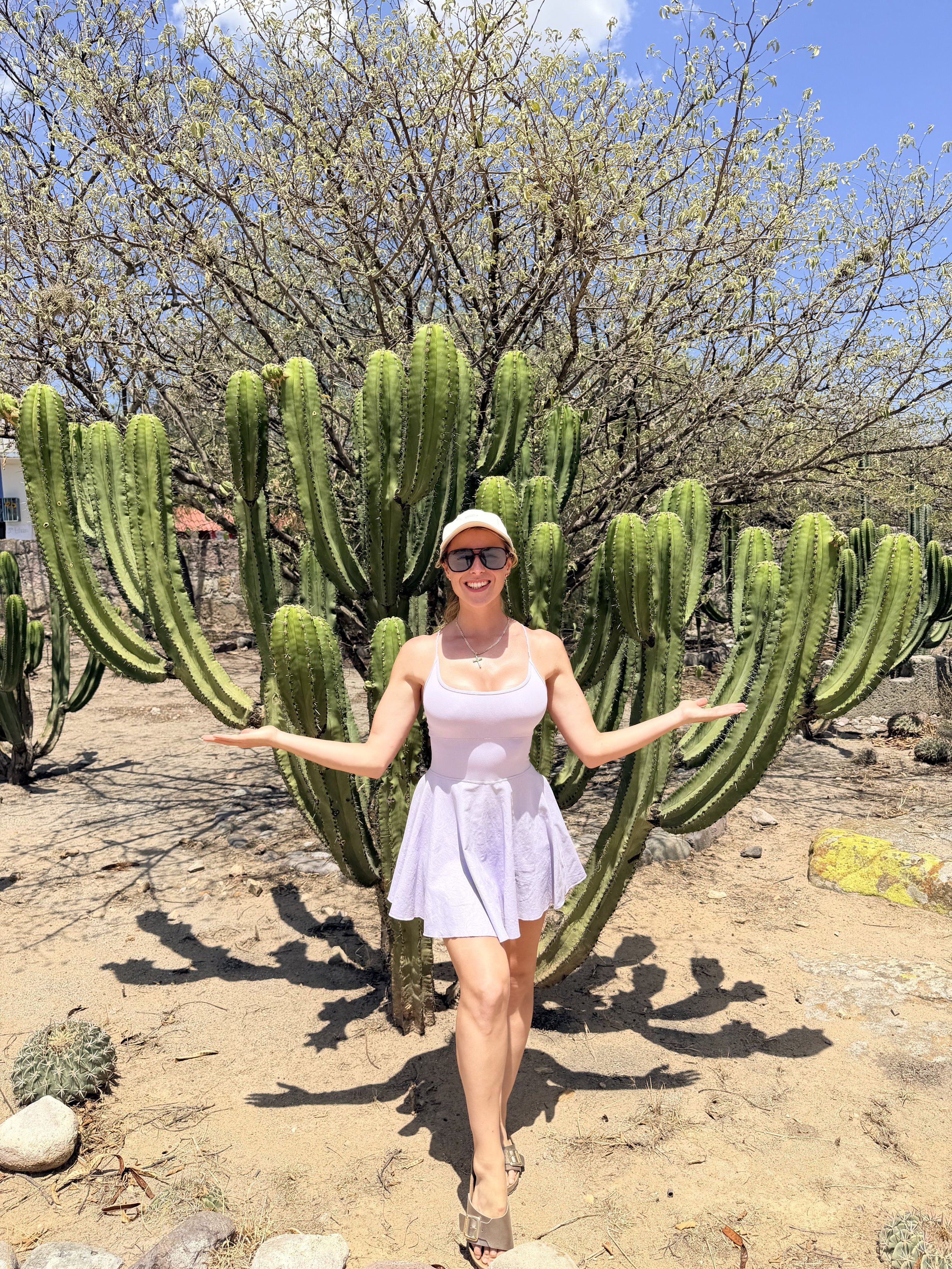 A woman wearing a white dress and sunglasses smiling with arms outstretched in front of tall cacti and a desert landscape under a clear blue sky.