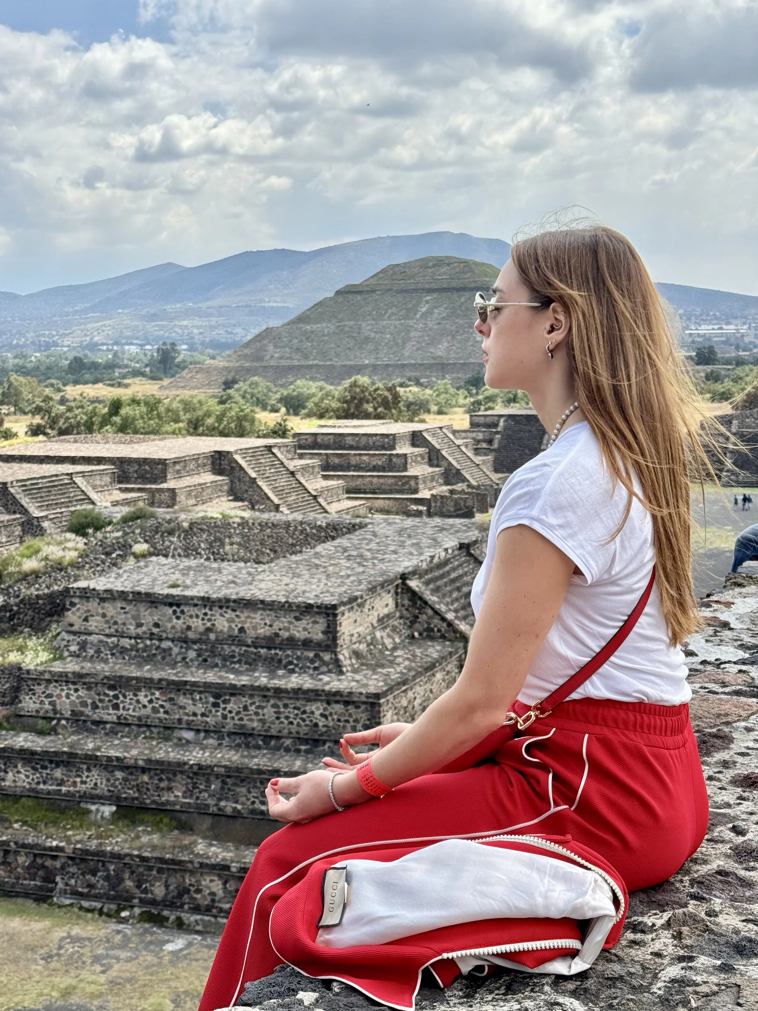 A woman with long red hair sitting cross-legged on ancient stone steps, facing to the right, with a pyramid and mountains in the background.