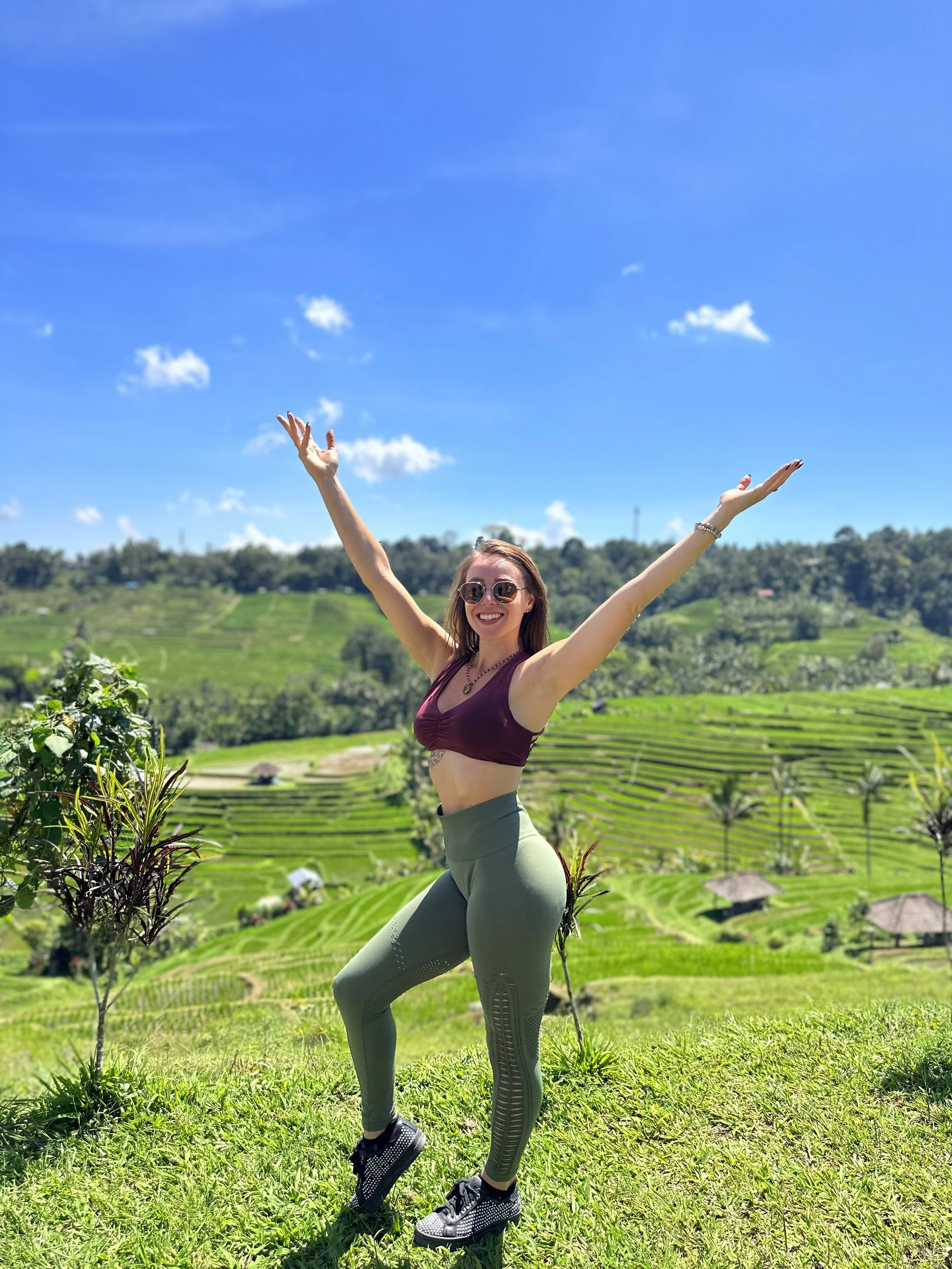 A woman with sunglasses and a purple sports bra smiling and raising her arms in a lush green outdoor landscape with terraced rice fields and blue sky.