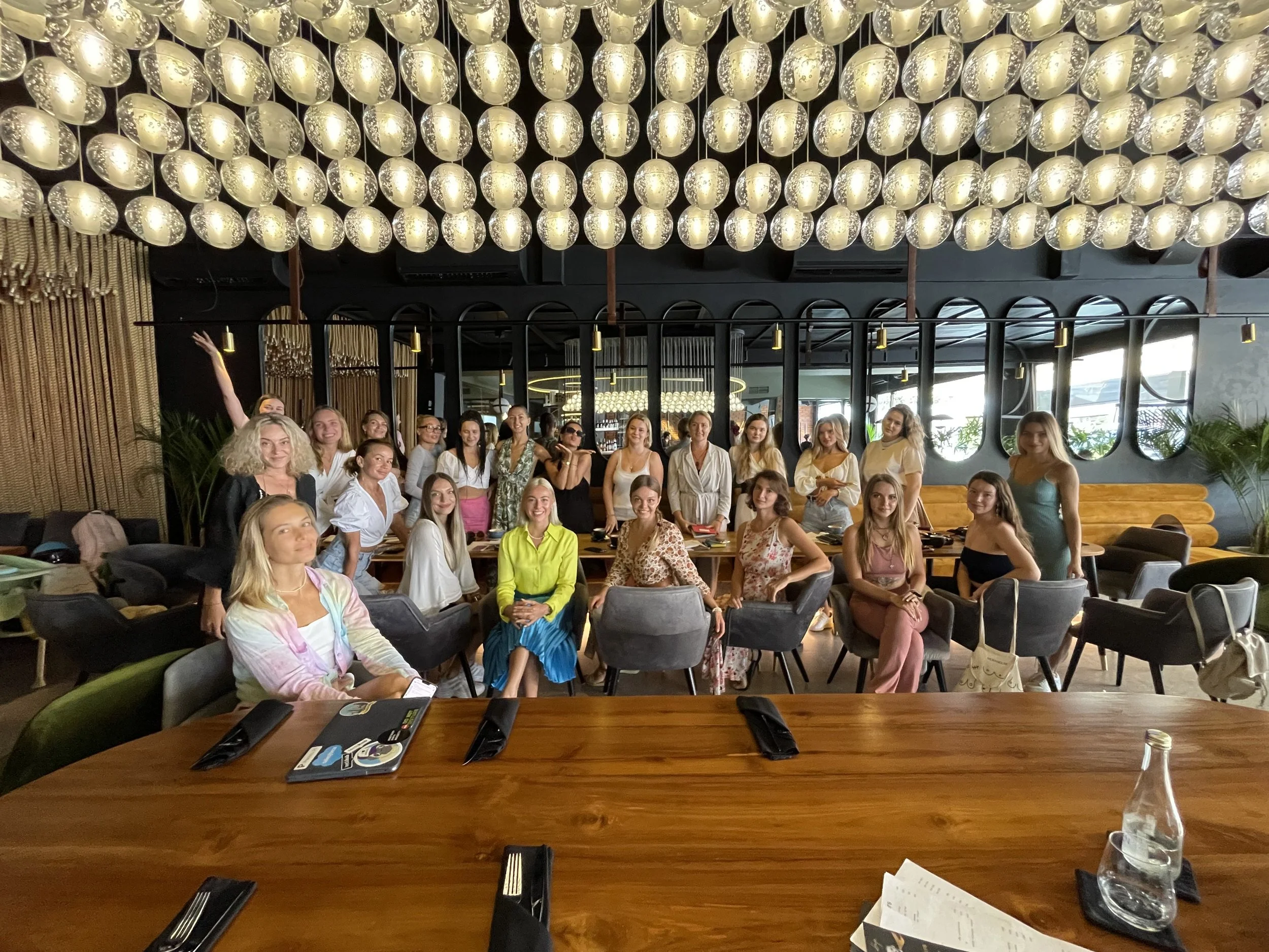 Group of women gathered in a restaurant or cafe with a modern, industrial design, illuminated by spherical hanging lights on the ceiling. The women are seated and standing around a large wooden table, smiling, with some looking at the camera. The setting includes some plants, mirrors, and a neutral color palette.