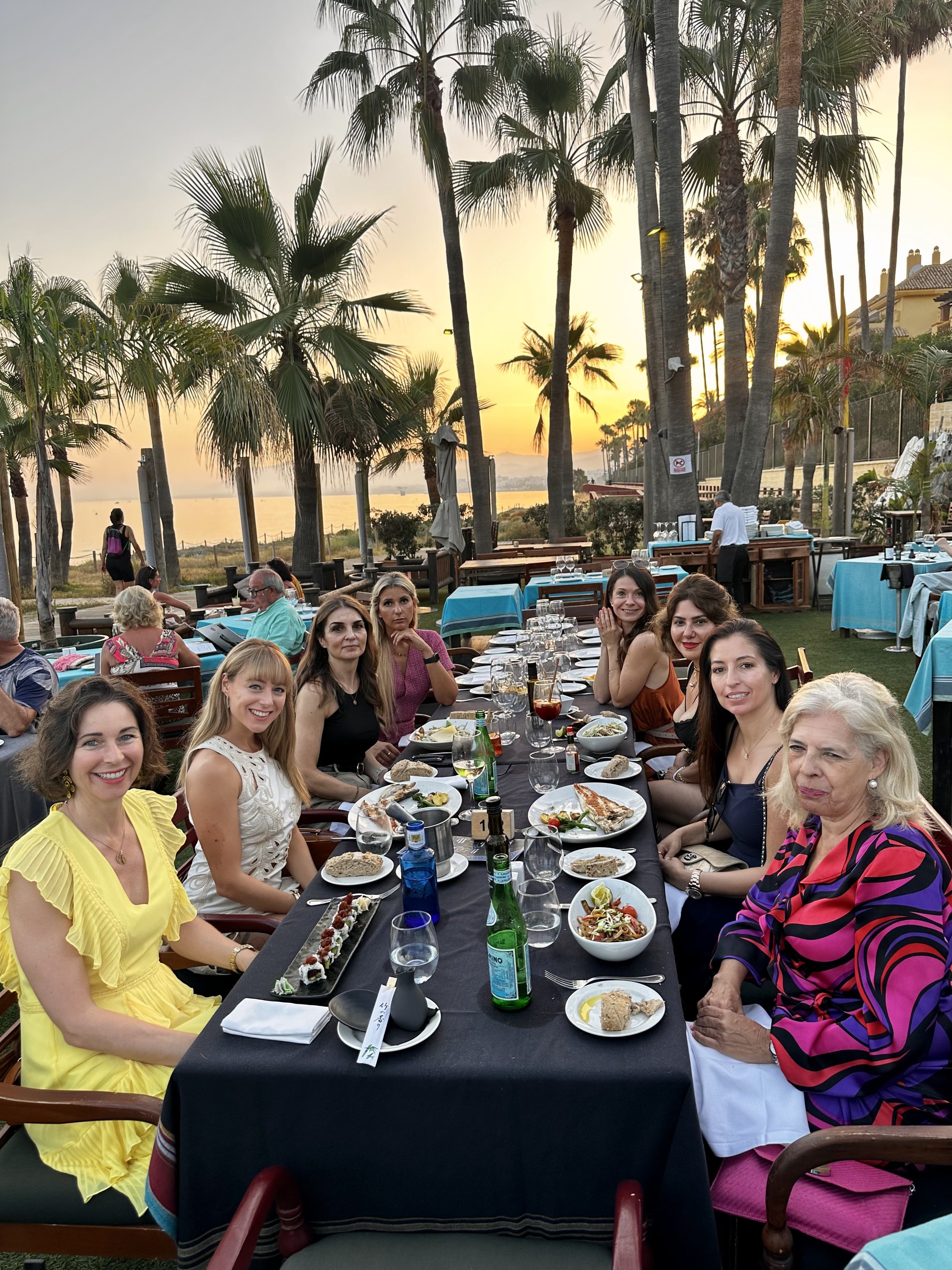 A group of women and men sitting at a long outdoor dining table overlooking a body of water during sunset. The table is set with plates of food, drinks, and chopsticks. Palm trees and a sunset sky are in the background.