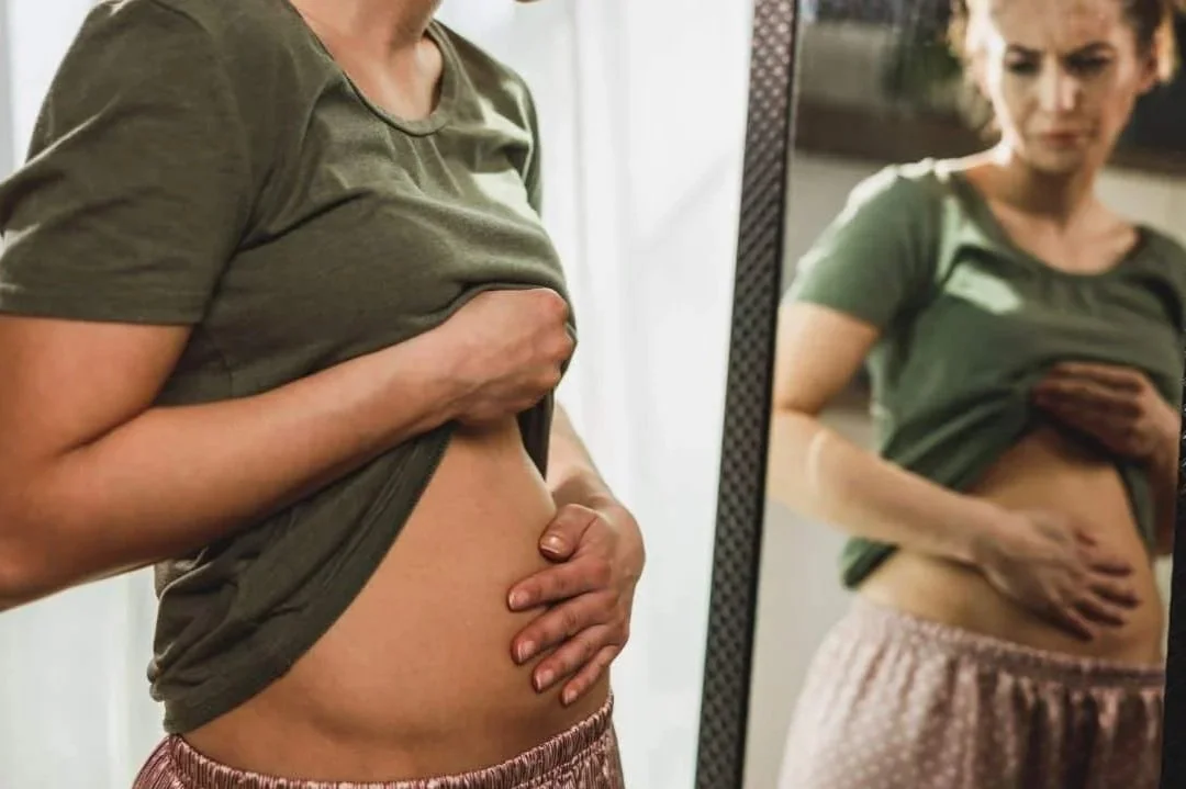 A woman is looking at her reflection in a mirror while checking her pregnant belly, wearing a green t-shirt and pink patterned pants.