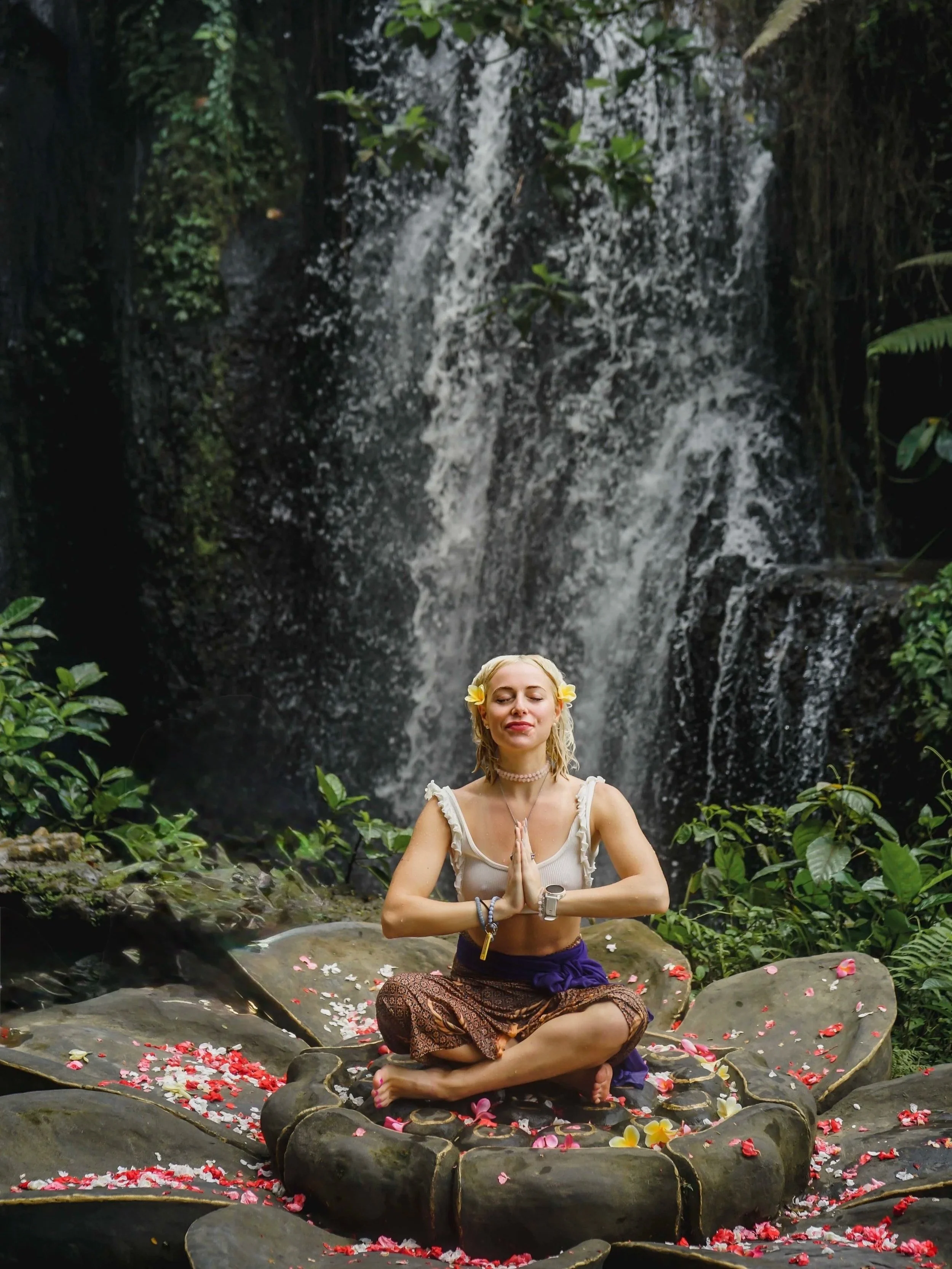 A woman sitting cross-legged on rocks in front of a waterfall, with her hands in prayer position, wearing a white top, patterned pants, and a flower in her hair, surrounded by flower petals.