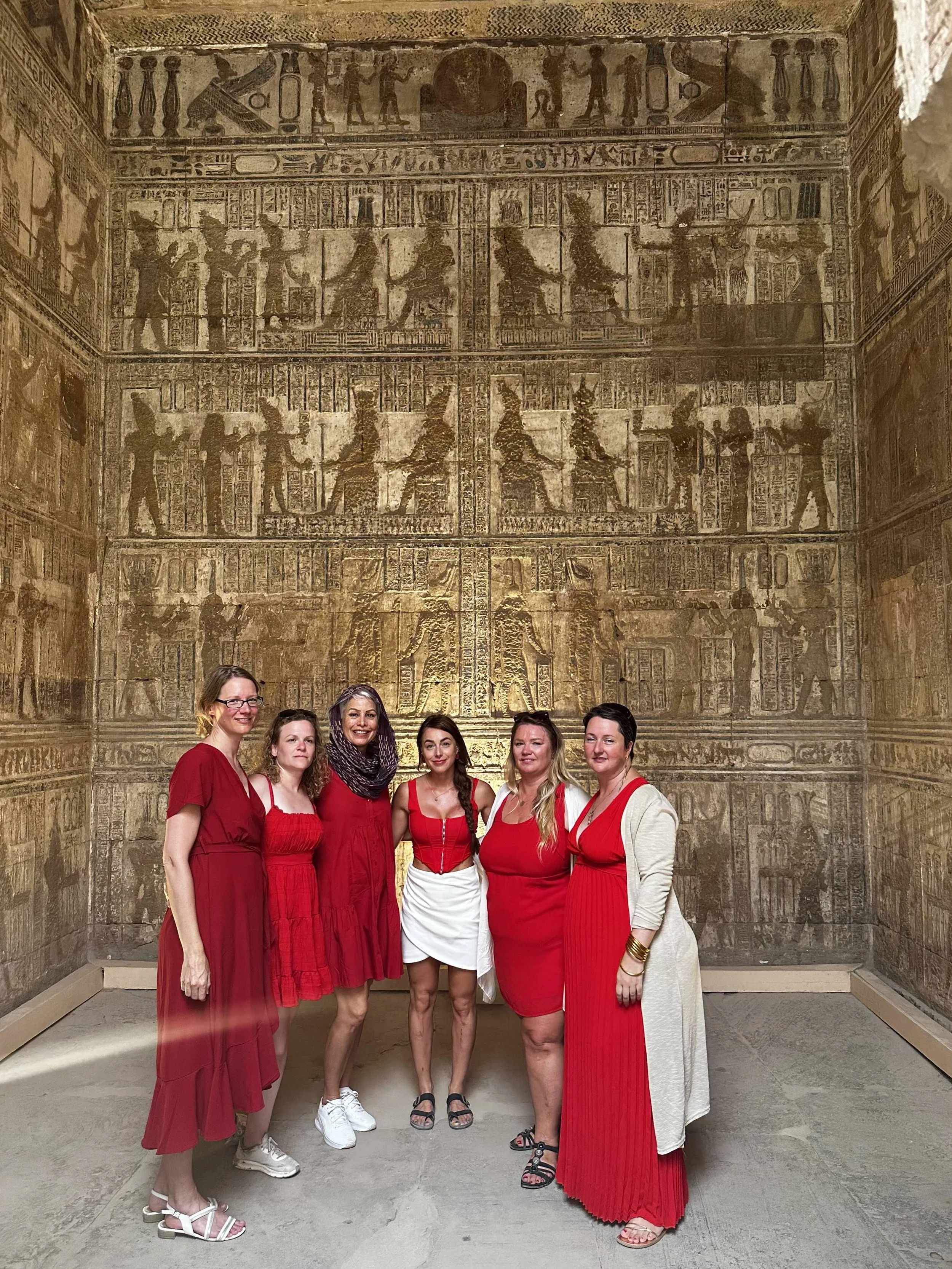 A group of six women dressed in red and white clothing standing in front of an ancient Egyptian wall with hieroglyphics and carvings.