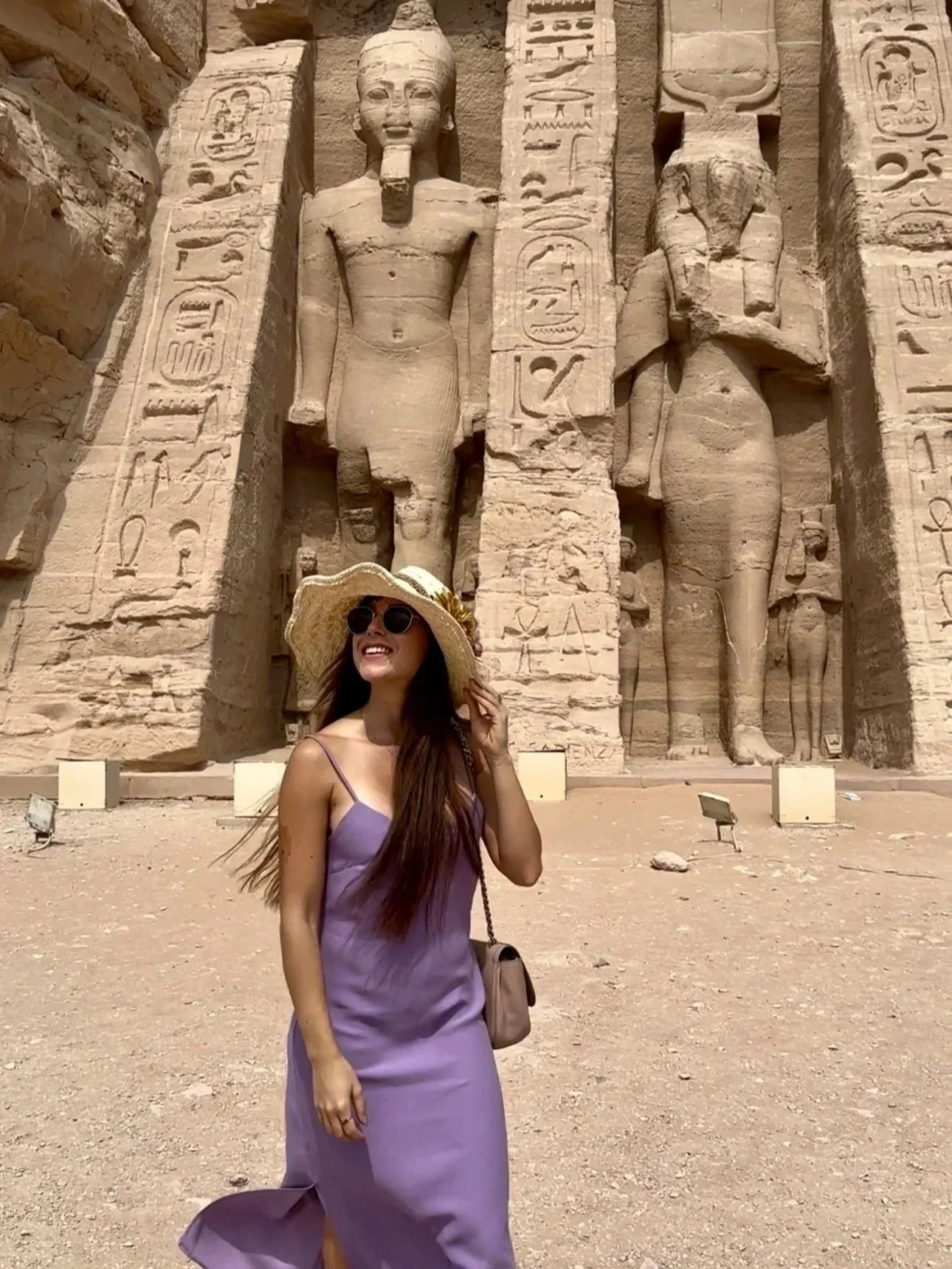 A woman in a purple dress and large sun hat smiling and standing in front of ancient Egyptian statues with hieroglyphics. The statues include a male figure with a headdress and a female figure with a tall crown. The setting appears to be a historical site in Egypt, possibly a temple or tomb.