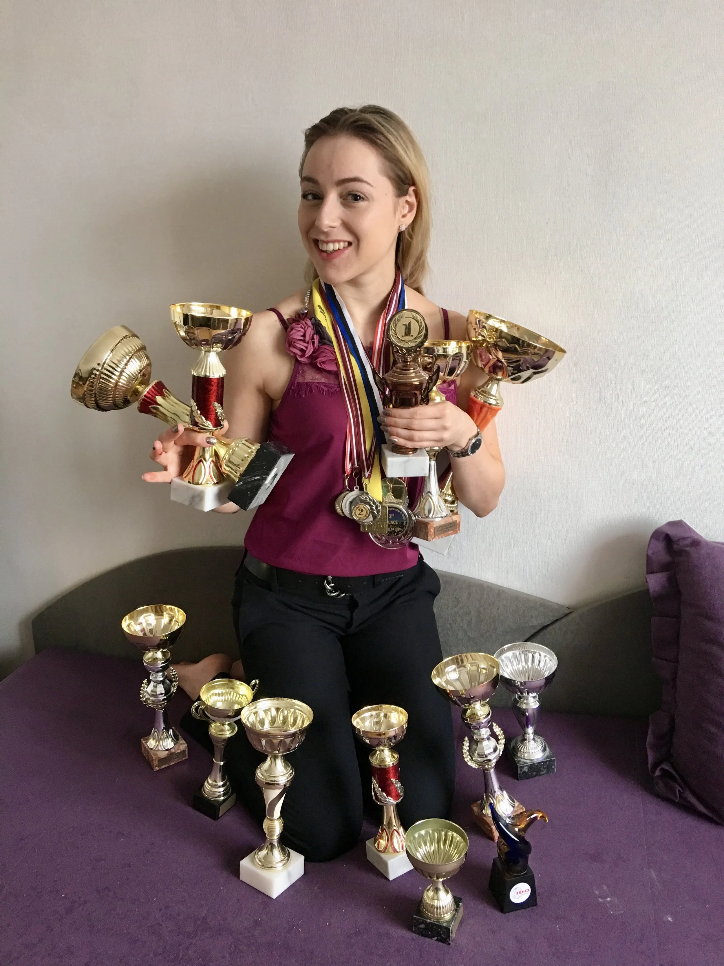 A woman with blonde hair smiling and holding multiple trophies and medals, standing in front of a plain wall. Several additional trophies are placed on a purple surface in front of her.