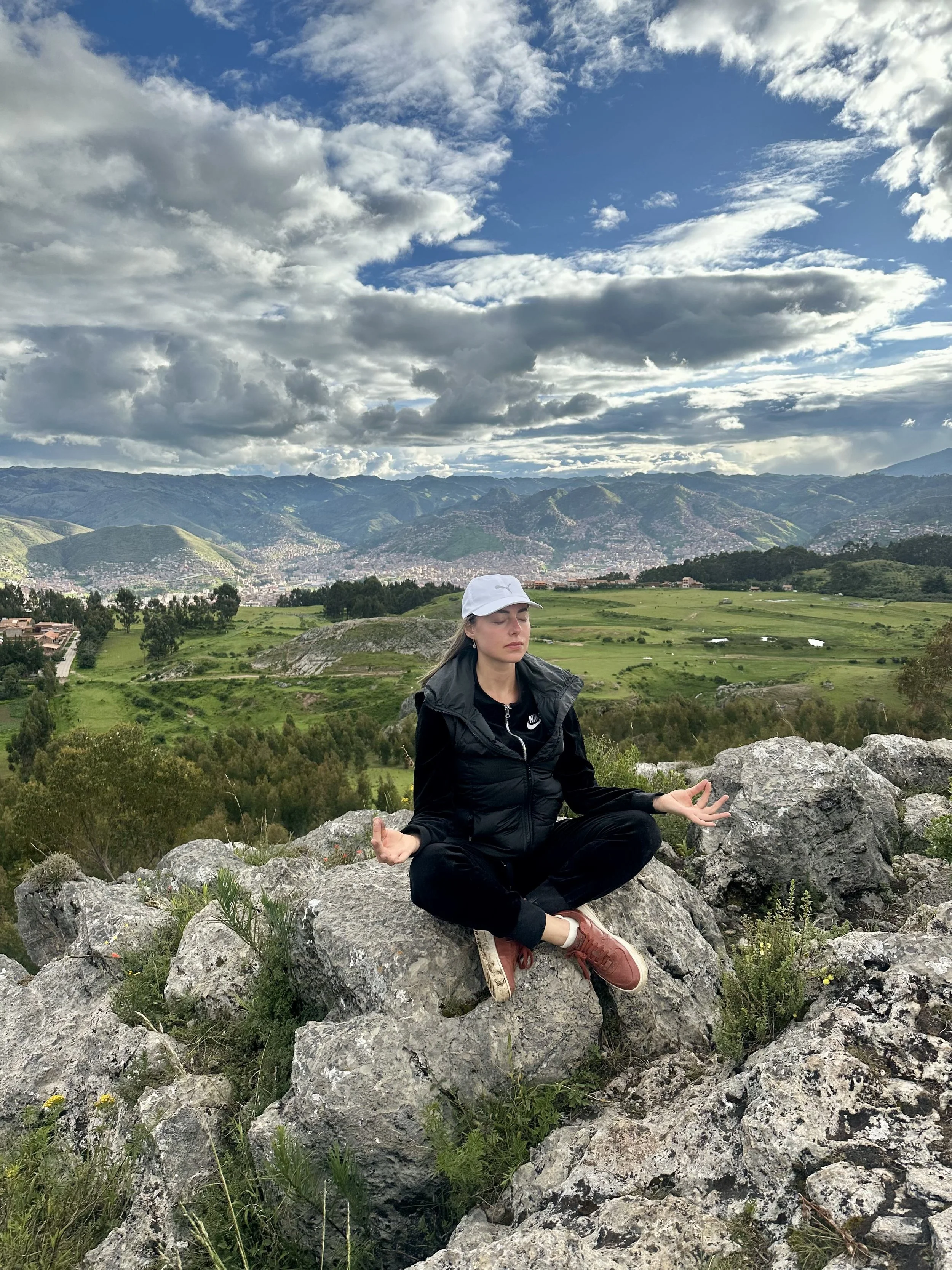 A woman practicing meditation in a seated yoga pose on rocks outdoors, with a scenic background of green hills, mountains, and a partly cloudy sky.