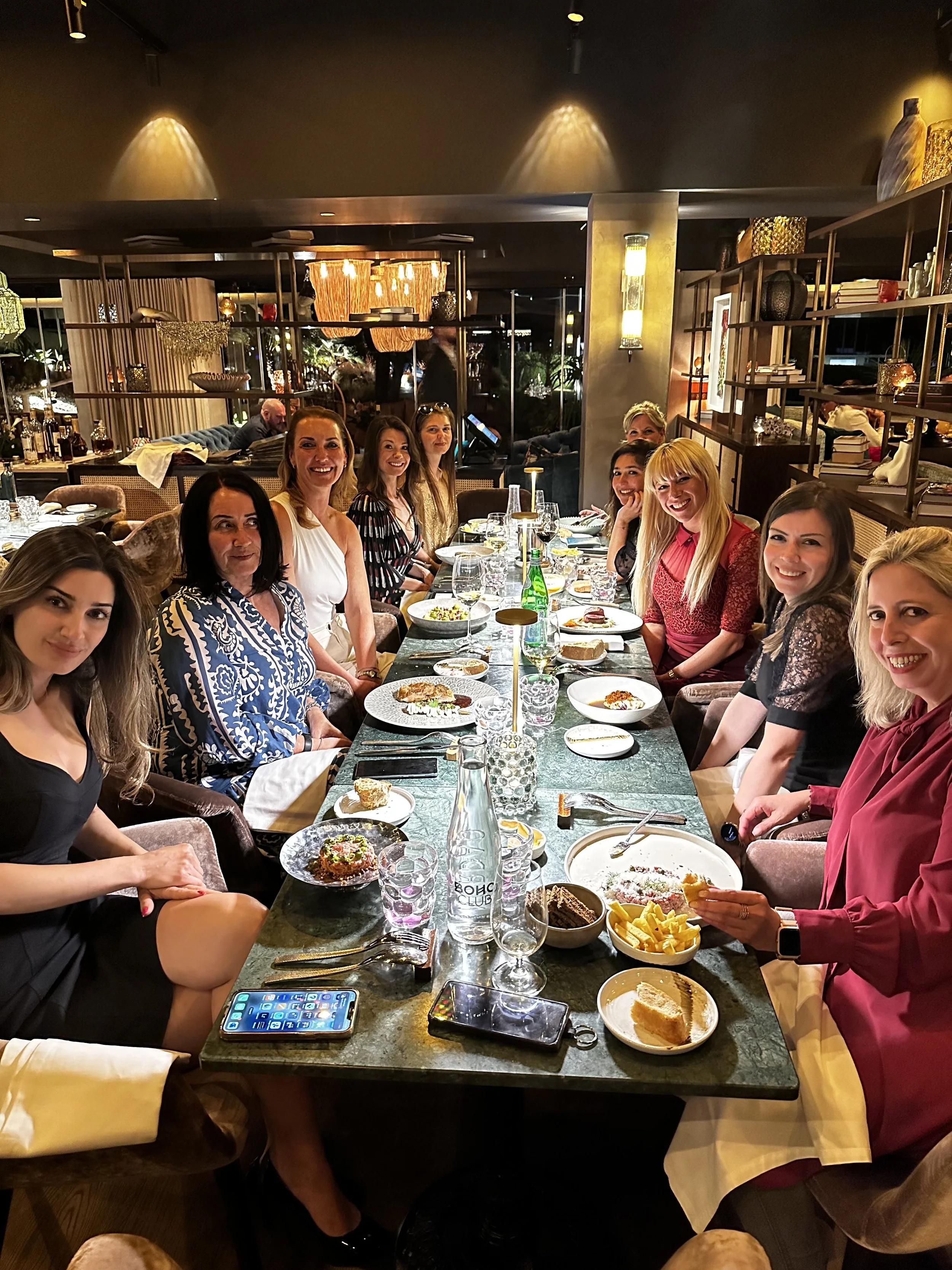 A group of women sitting at a long dining table in a restaurant, smiling and enjoying a meal with various dishes and beverages, with warm lighting and decorative shelves in the background.