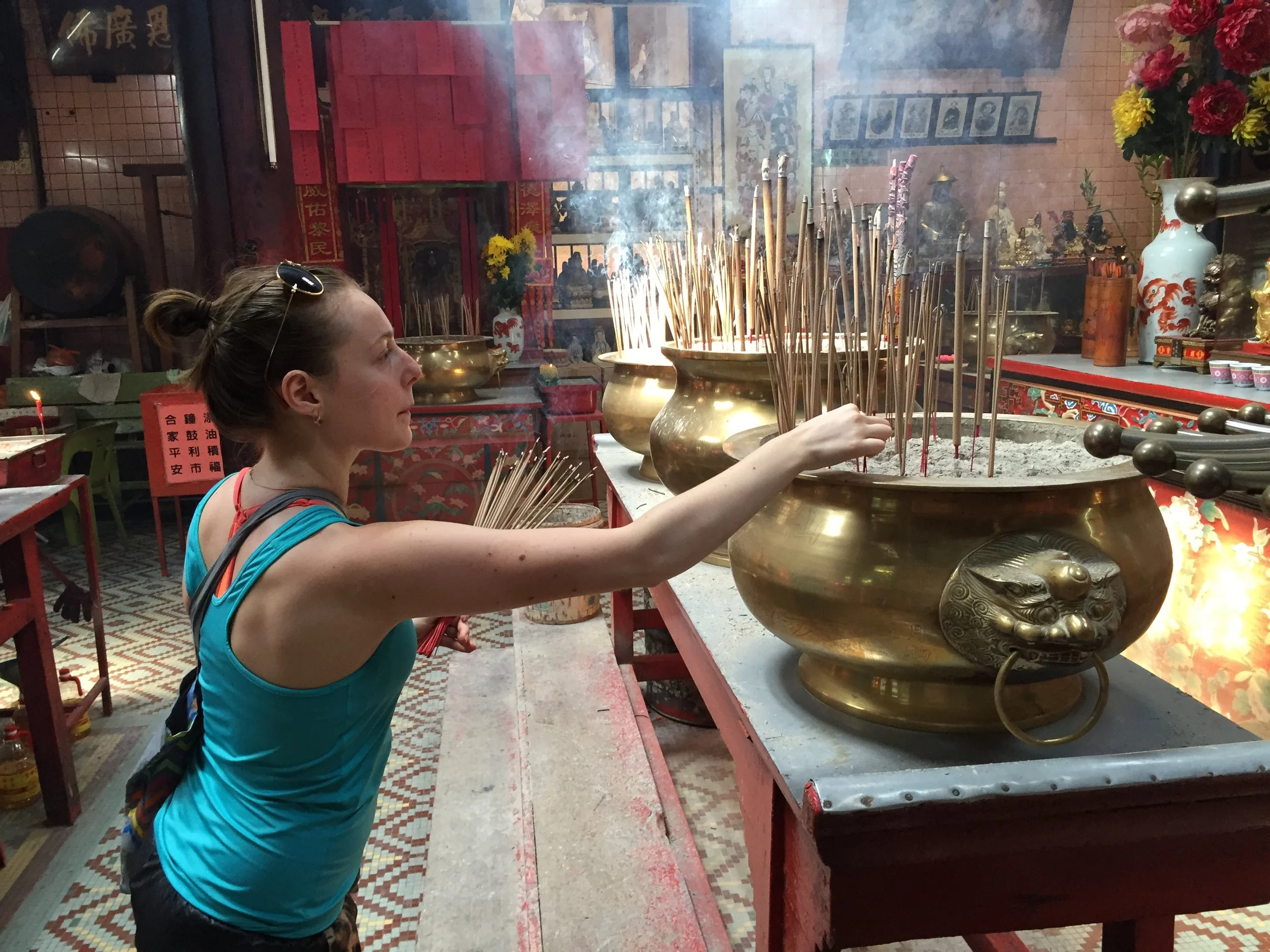 A woman with sunglasses on her head wearing a blue top holds incense sticks in a temple. She is lighting incense at a large incense burner filled with ash and many incense sticks, with smoke rising from them. The background features traditional Chinese decorations, vases, shrine statues, and framed photographs.