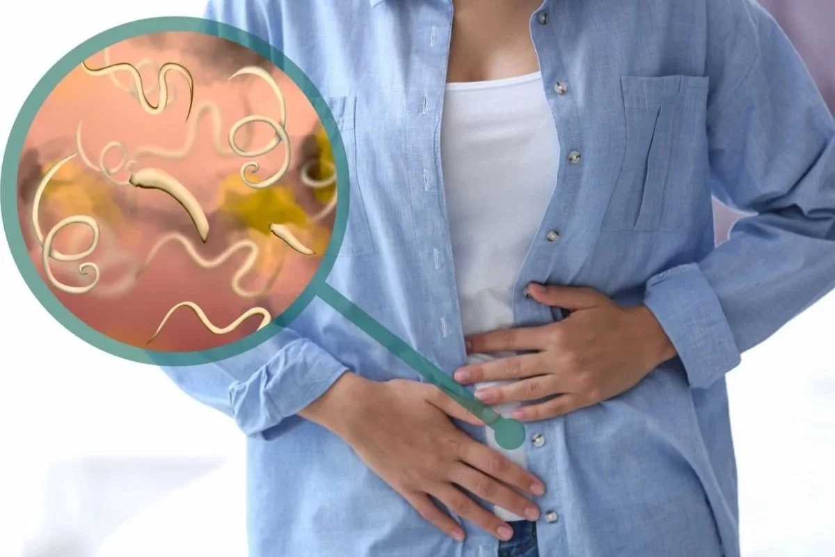 A woman with a hand on her stomach, wearing a light blue denim shirt and white T-shirt, with a graphic illustrating a close-up of intestinal worms inside the intestine.