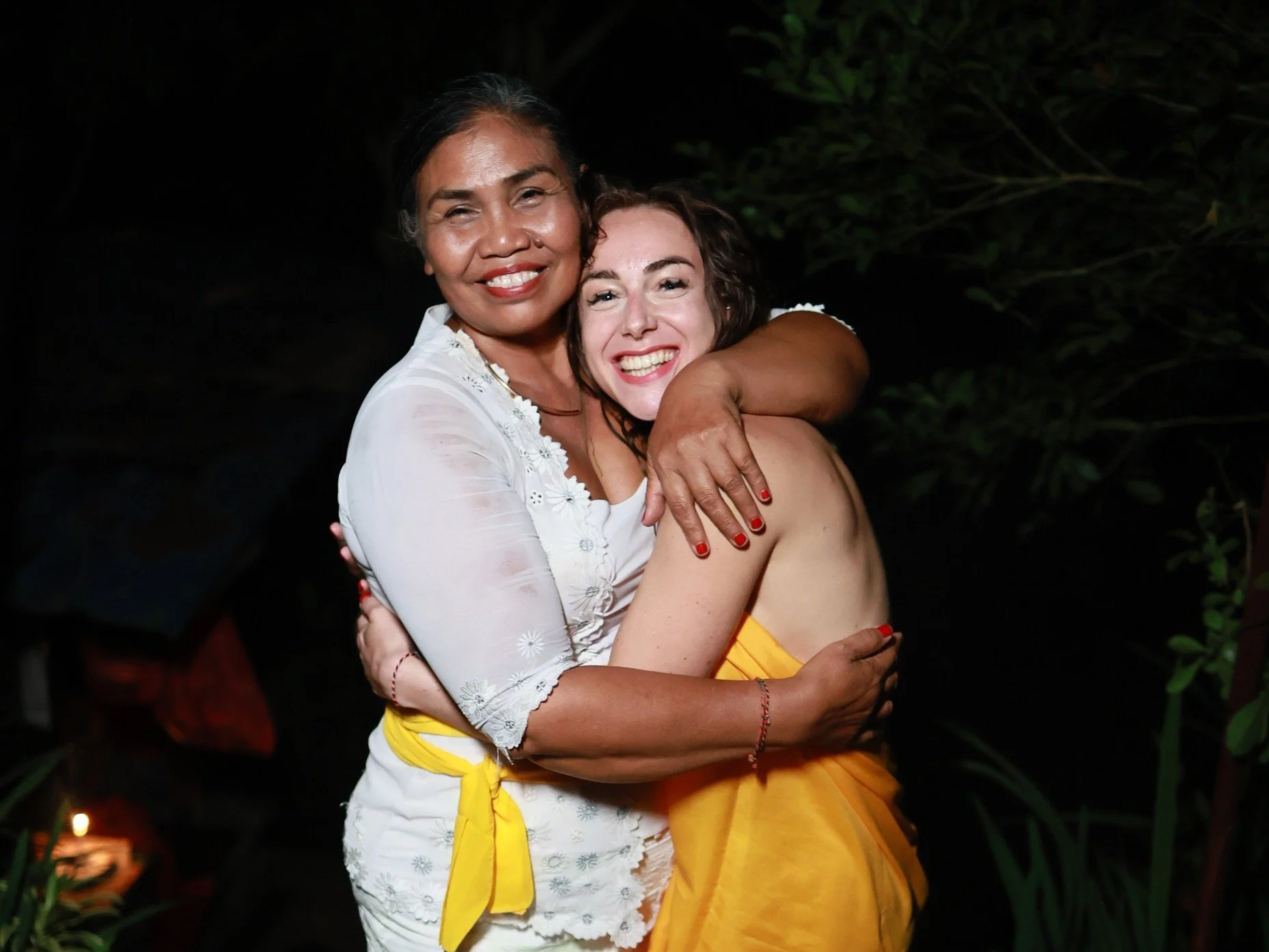 Two women embracing and smiling at night, with a dark background and greenery around them.