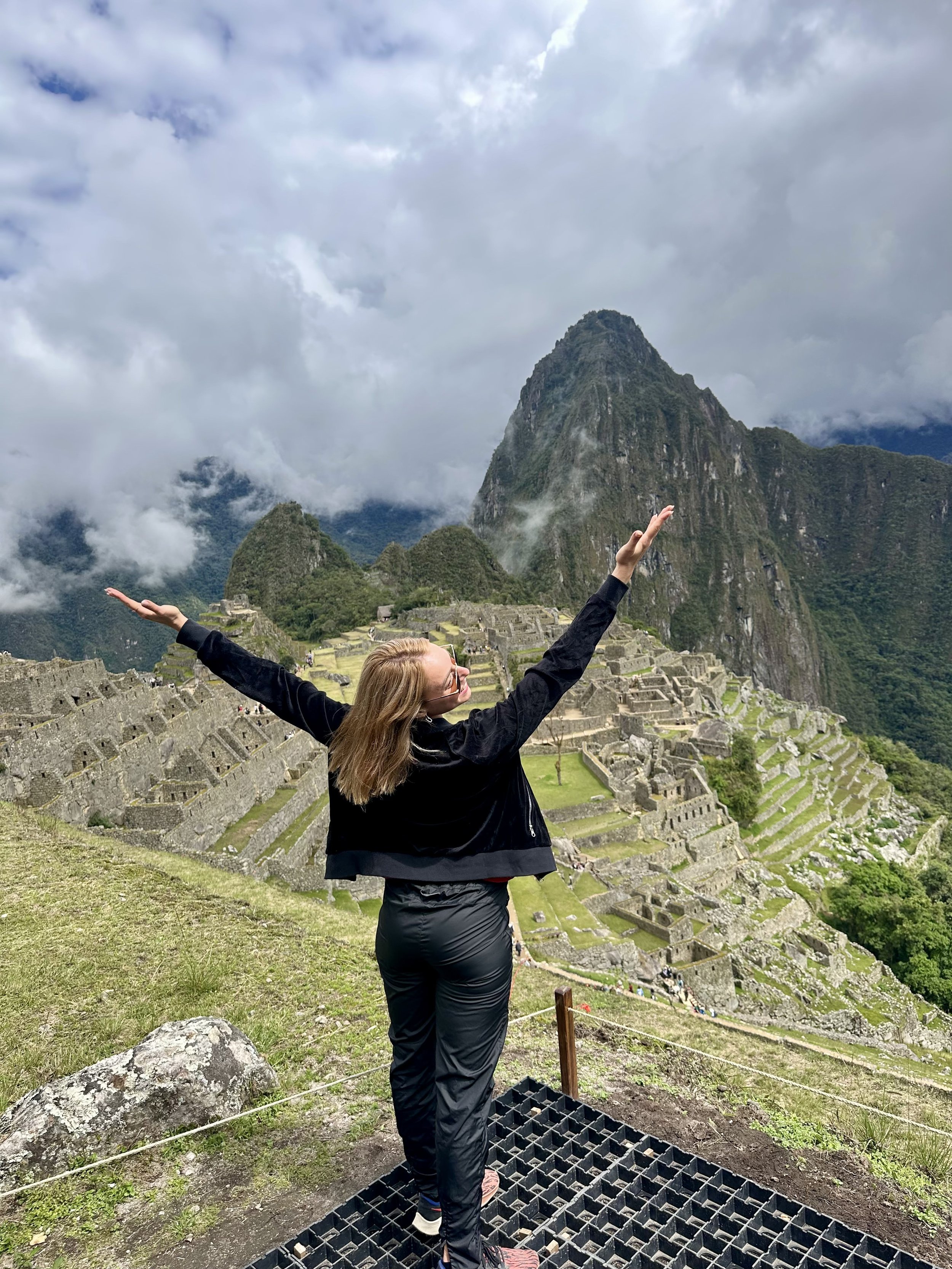 A woman stands with arms raised in front of Machu Picchu ruins in Peru, with Machu Picchu mountain and clouds in the background.