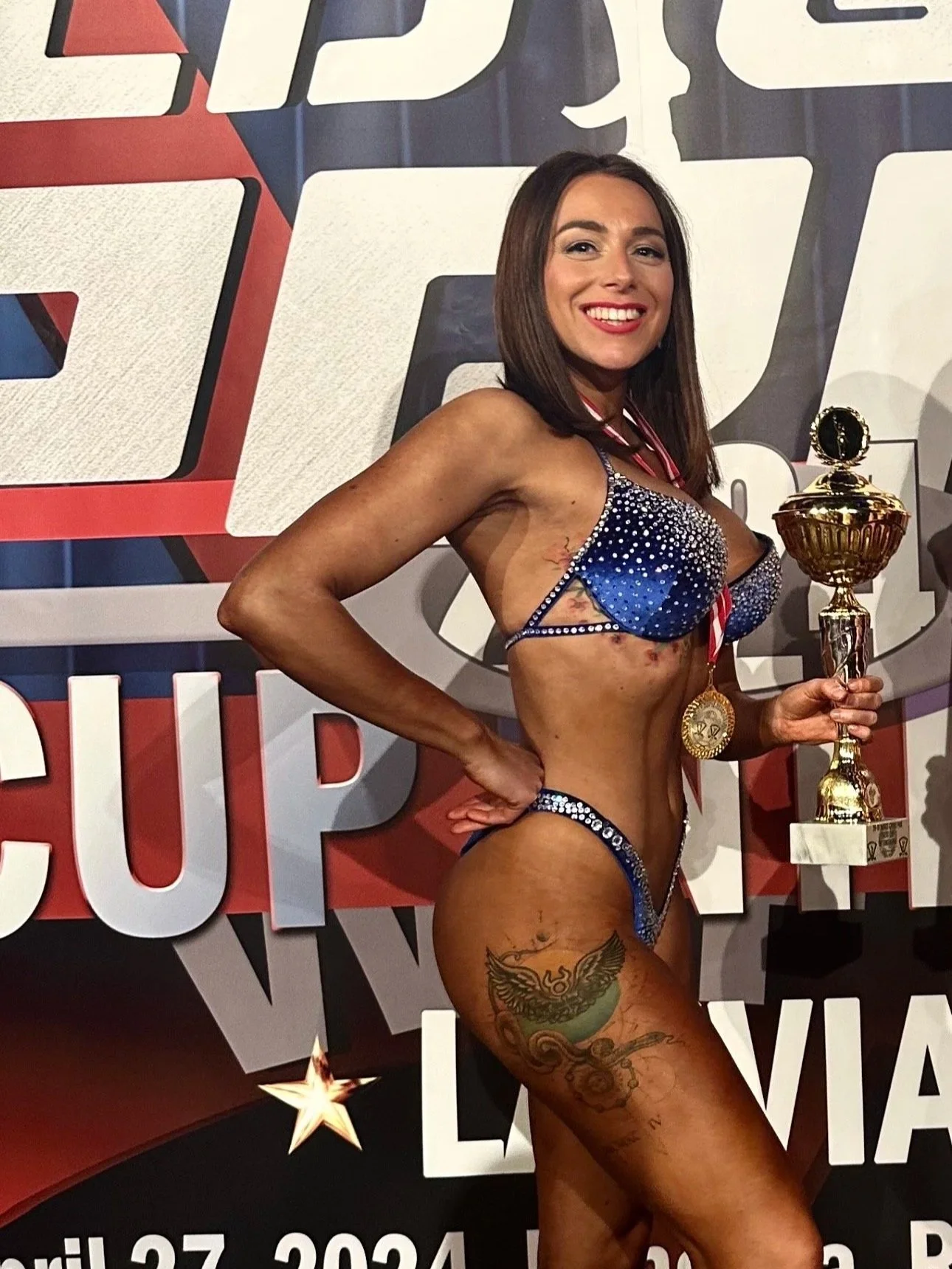 A woman in a shiny blue bikini with rhinestones smiles and poses with a golden trophy in front of a backdrop with large stylized letters and stars.