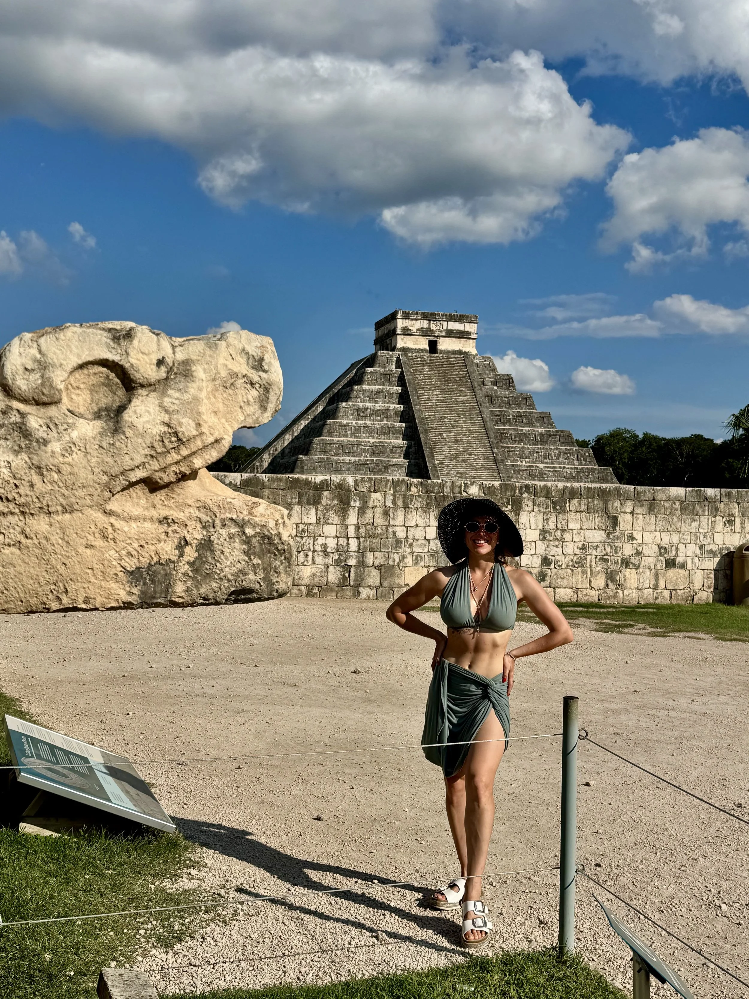 Woman in a green dress and large black hat standing in front of ancient Mayan pyramid at Chichen Itza, Mexico, with a dinosaur stone head sculpture in the foreground and informational signs on the ground.