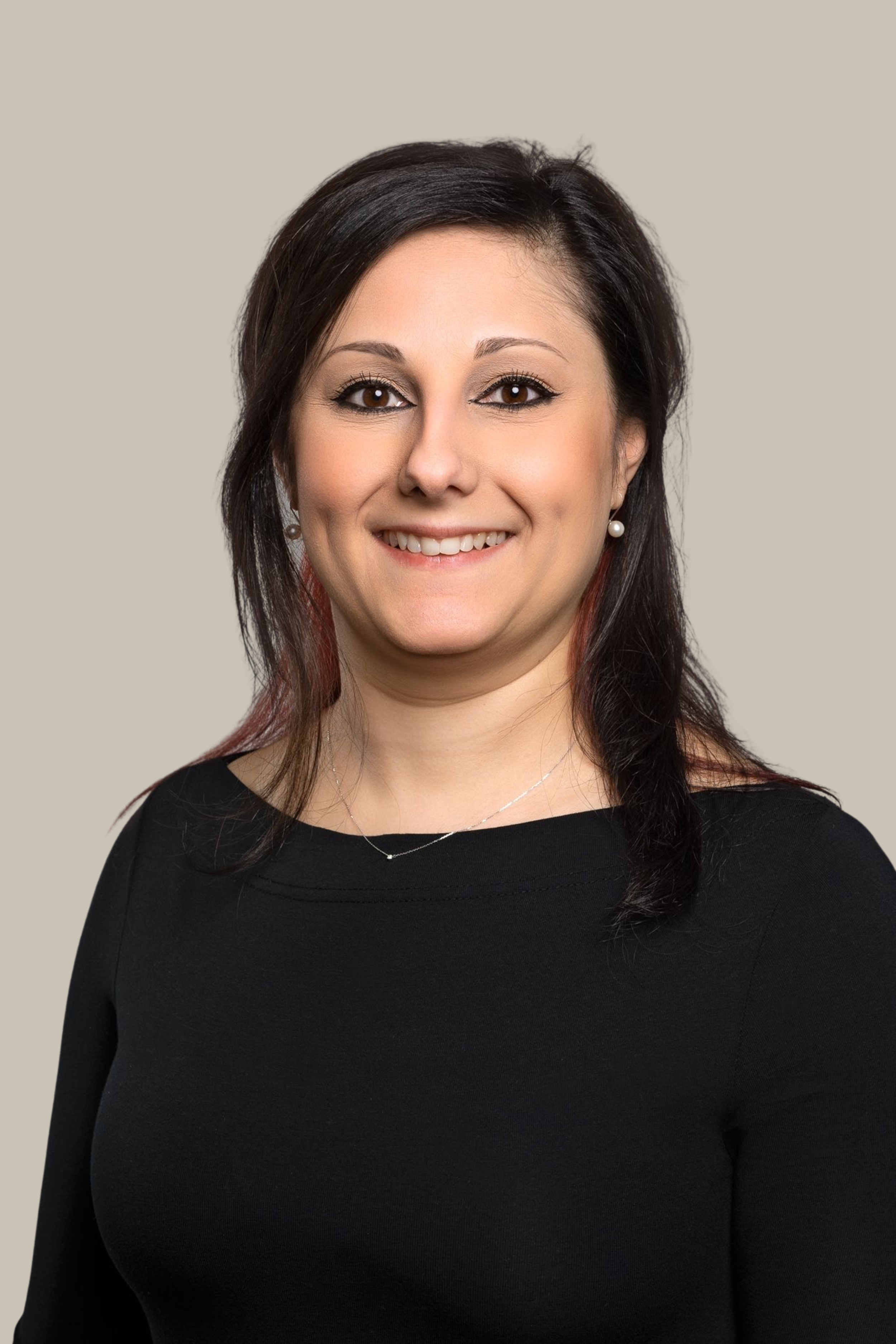 A woman with dark brown hair, brown eyes, and wearing pearl earrings and a black top, smiling against a plain light beige background.