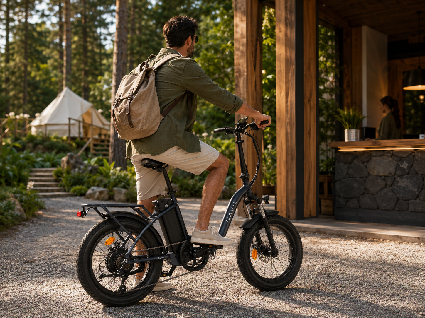 Un homme en short et chemise descend d'un vélo électrique devant un bâtiment en bois dans un cadre forestier. Un tipi est visible en arrière-plan.