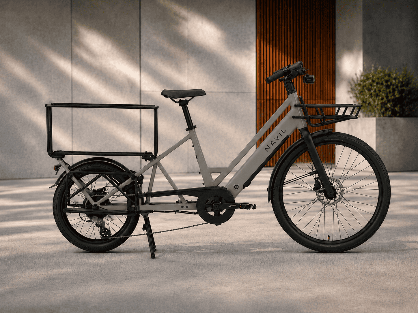 Vélo électrique gris avec porte-bagages, stationné sur un sol en béton, avec un mur en pierre derrière et un meuble en bois à droite, une plante dans un grand pot en béton à l'arrière