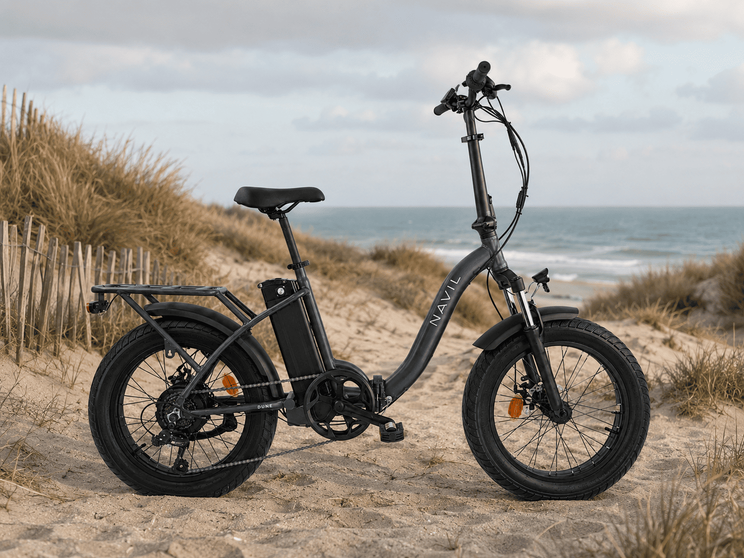 Vélo électrique noir sur une plage avec dunes et mer en arrière-plan.