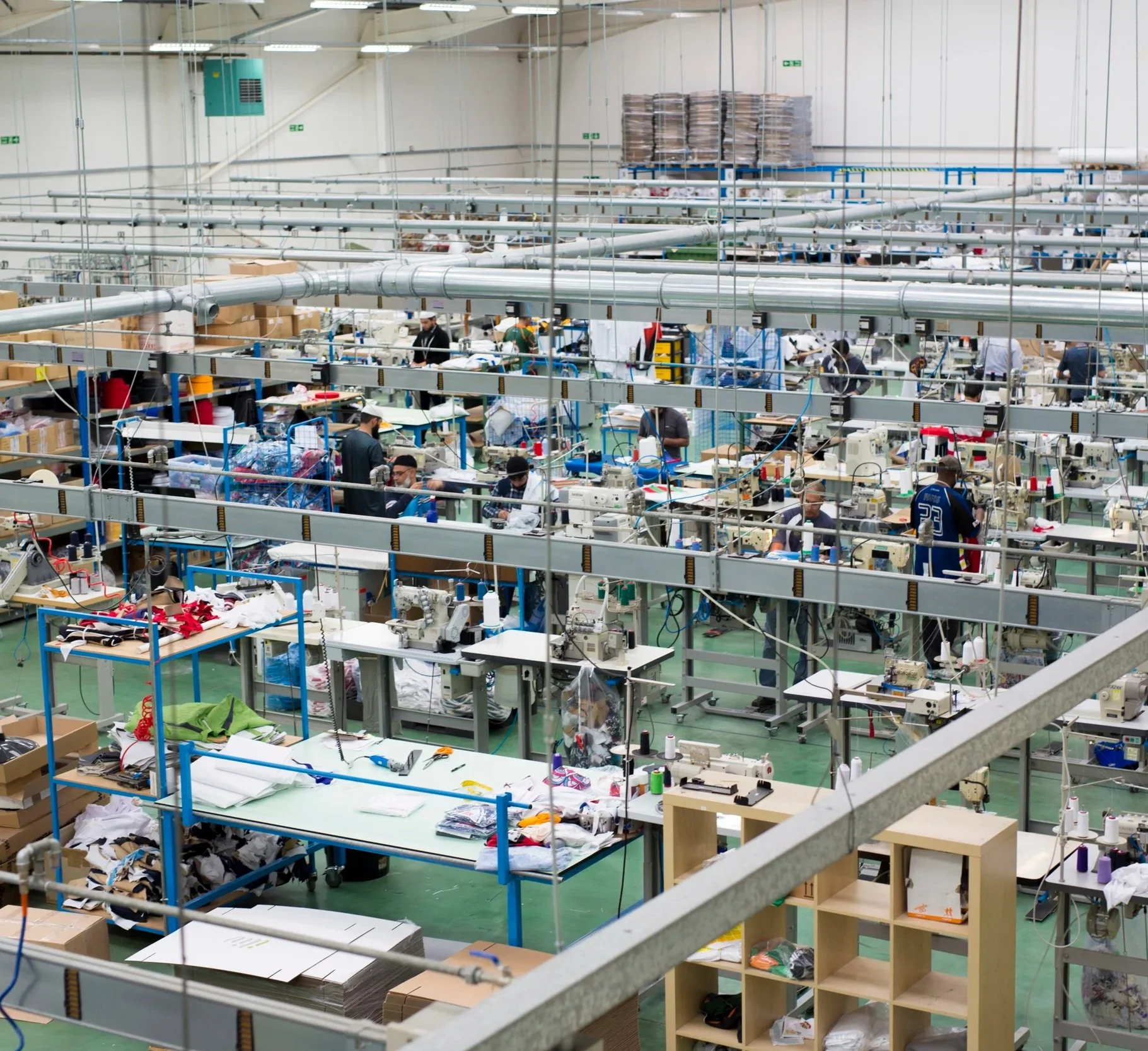 An expansive factory or warehouse with rows of workstations, sewing machines, and fabric or material pieces, with workers engaged in textile and garment production.