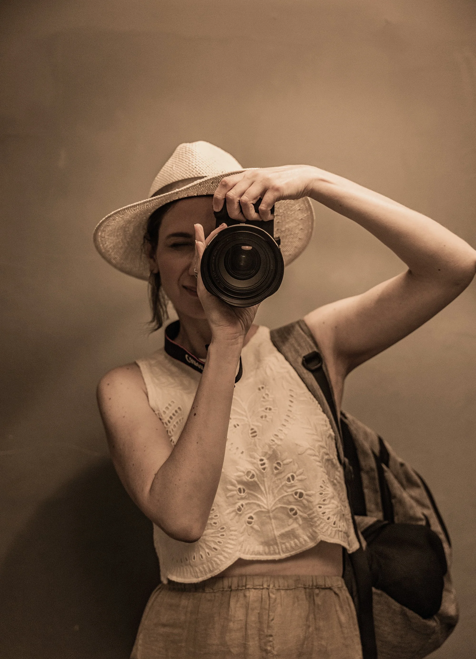 A woman takes a selfie in front of a mirror, wearing a straw hat and a beige lace top, with a backpack on her shoulder.