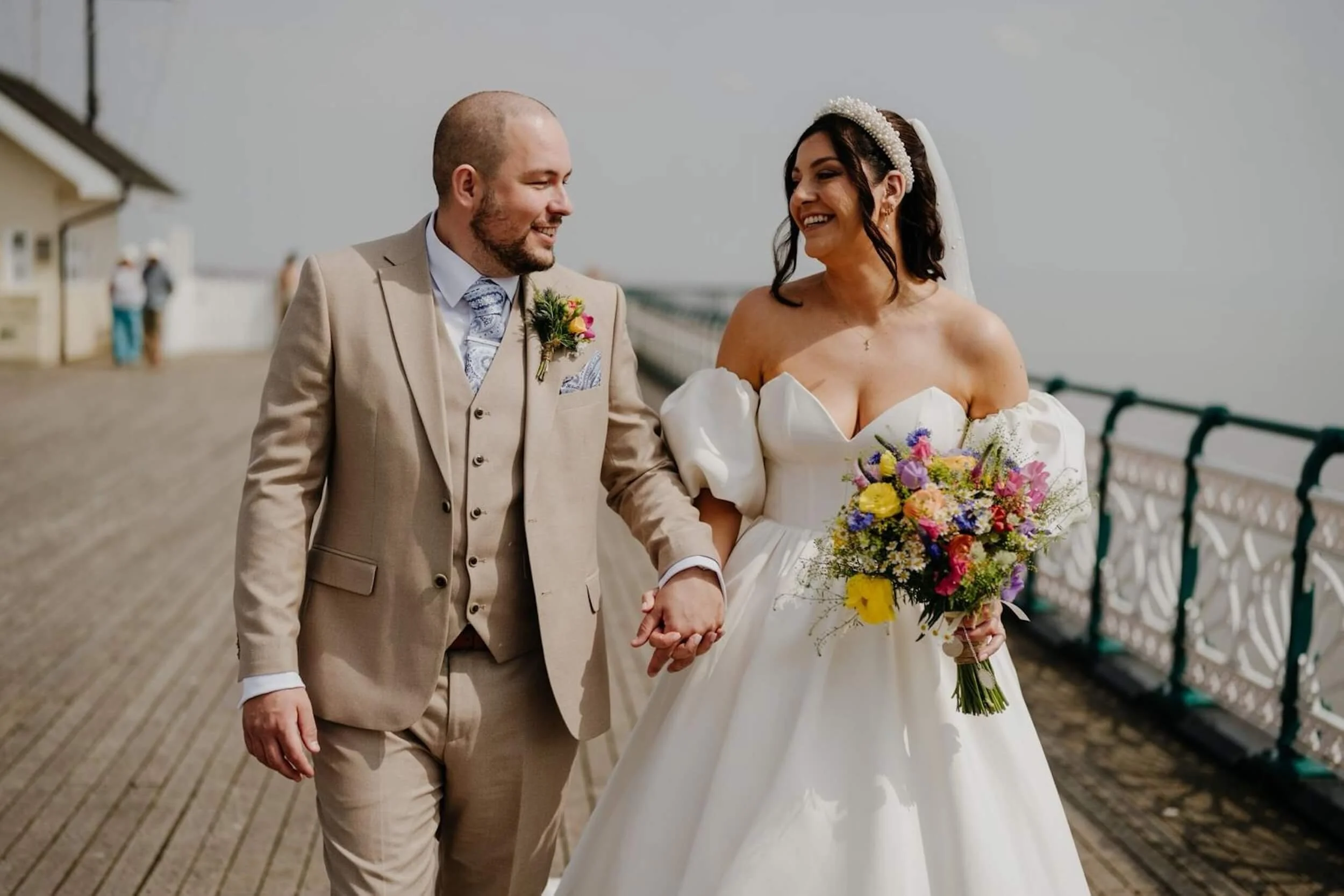 A newlywed couple walking hand in hand outdoors on a pier, smiling and looking at each other, with a woman in a white wedding dress holding a colourful meadow-style bouquet and a man in a beige suit with a matching buttonhole..