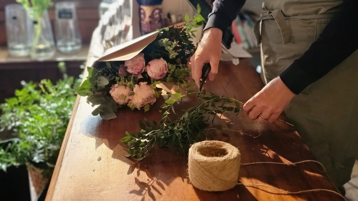 Person wrapping a bouquet of pink flowers and green foliage with brown paper on a wooden table. 