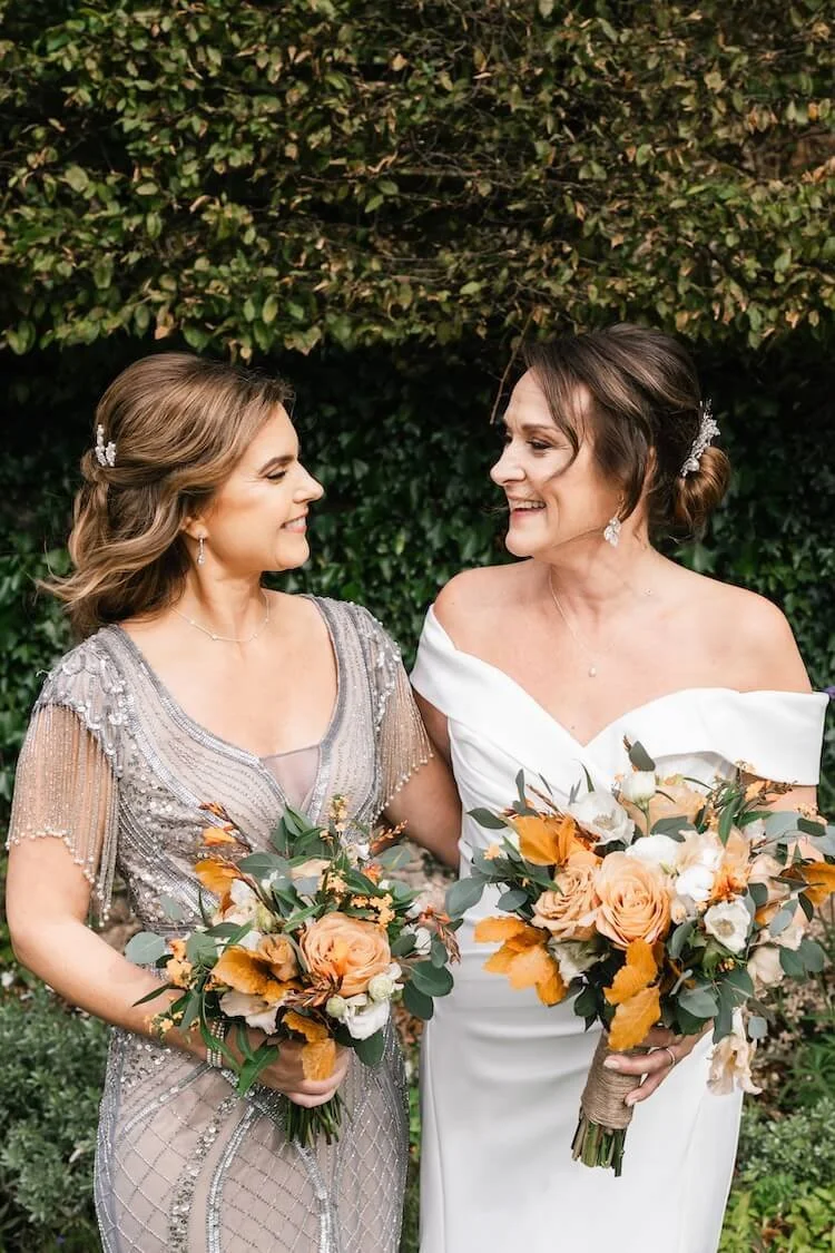 Bride and bridesmaid smiling at each other outdoors, holding bouquets featuring seasonal autumnal blooms, bound with natural twine