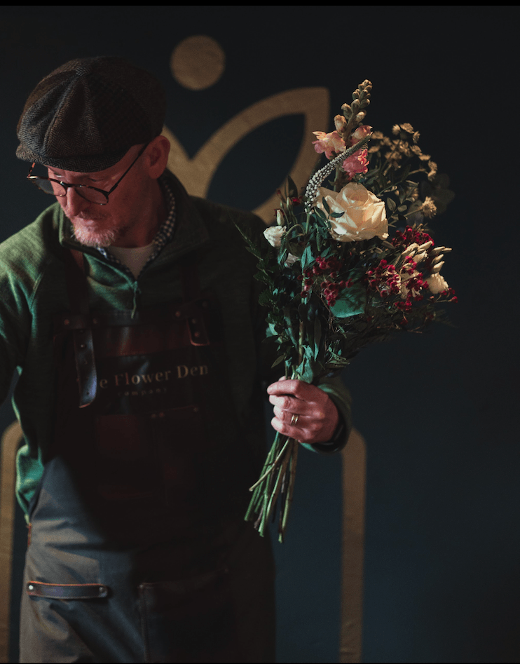 A man wearing glasses, a cap, and a dark apron is holding a bouquet of flowers.