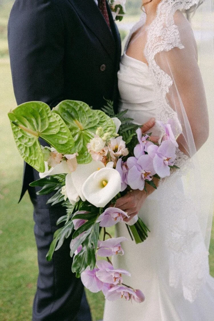 A bride and groom holding a large bouquet of flowers on their wedding day, with bride wearing a lace wedding dress and veil, and groom in a dark suit.